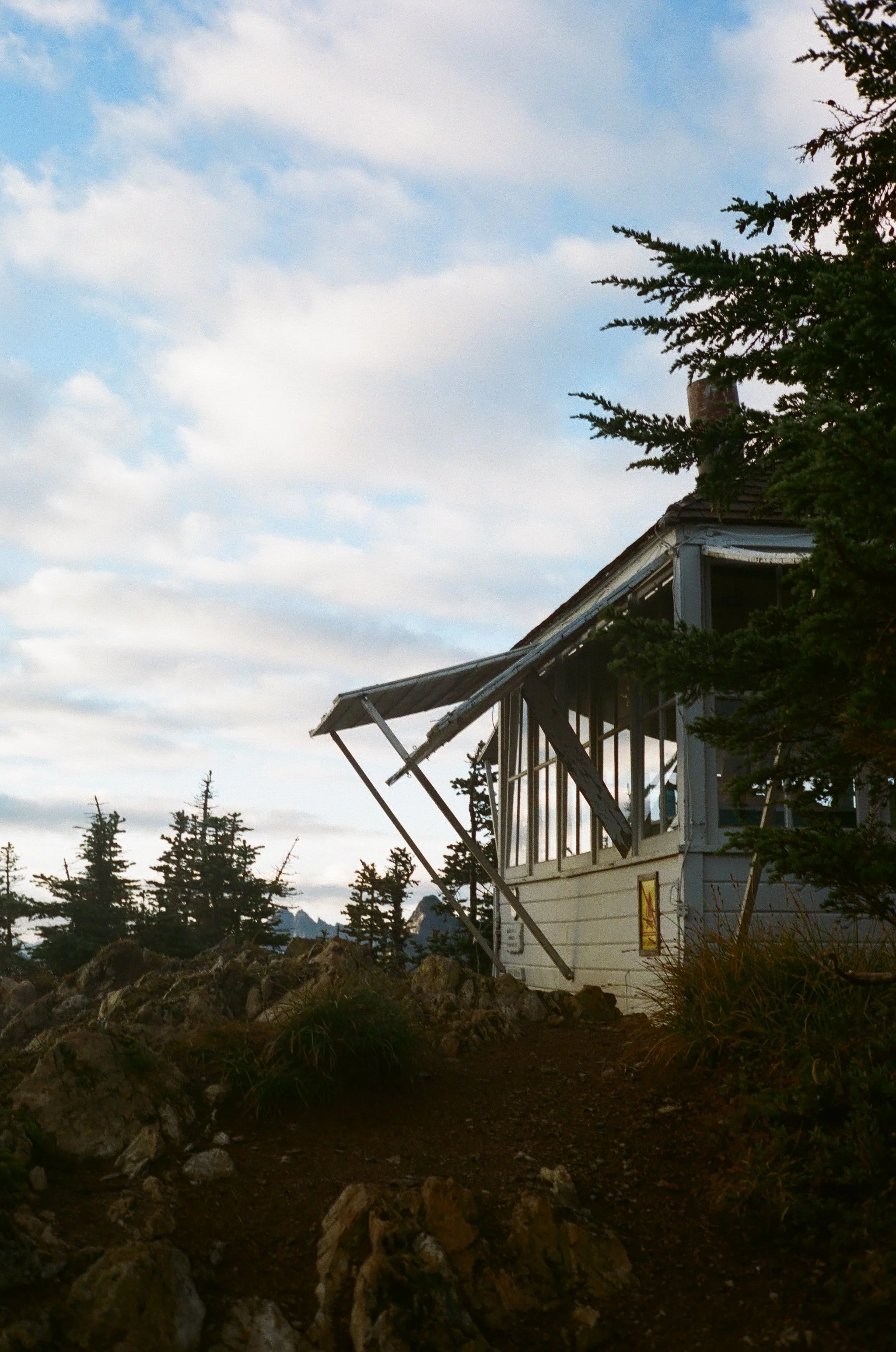 A small, weathered house on a rocky trail in a forested area, with a cloudy sky overhead.