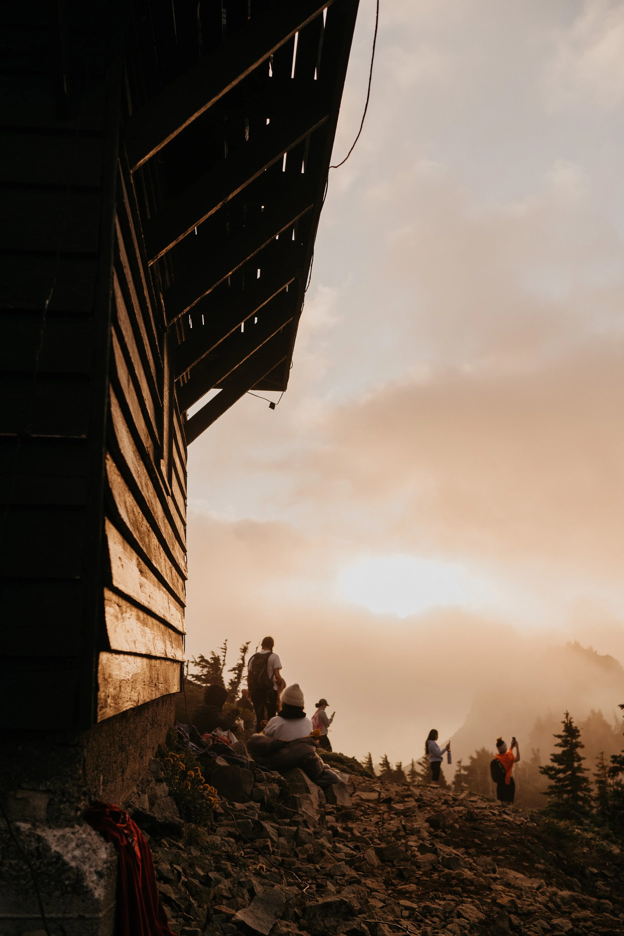 Group of people resting and taking photos near a mountain ridge during sunset, with a wooden structure on the left and misty mountains in the background.