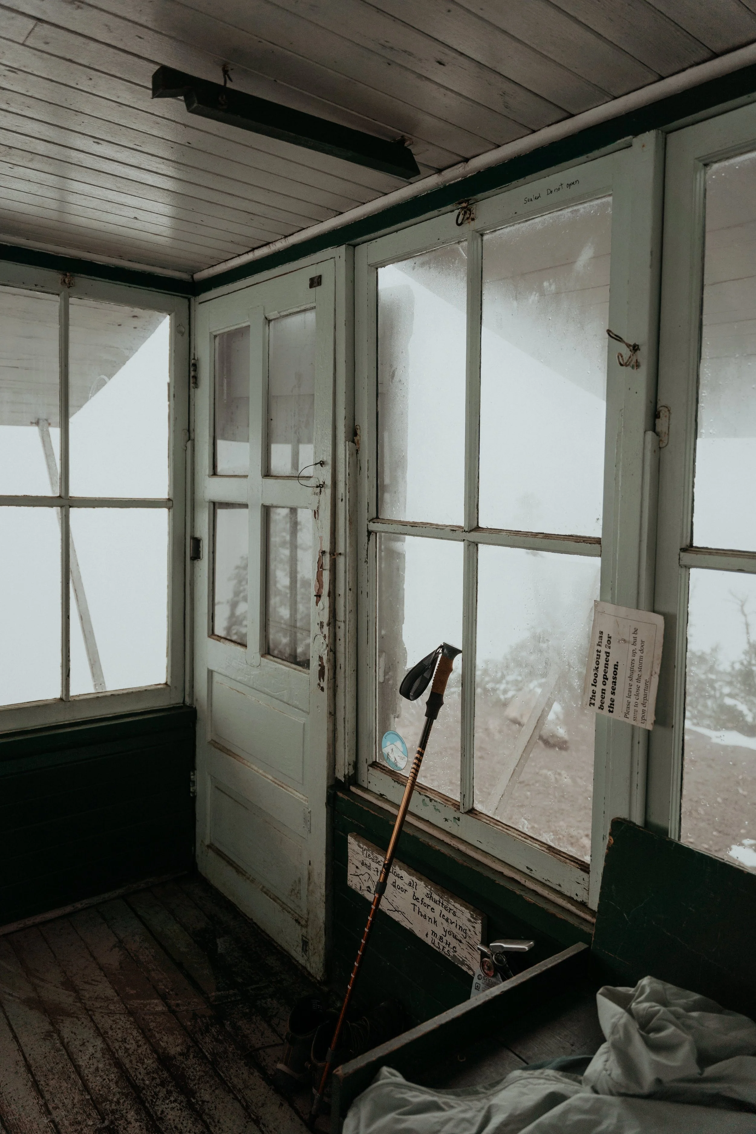 A rustic enclosed porch with weathered wooden flooring and window panes. A white wooden door with multiple glass panels and signs about the season and door usage. A golf club and shoes are seen in the corner, and foggy outdoor scenery is visible thro