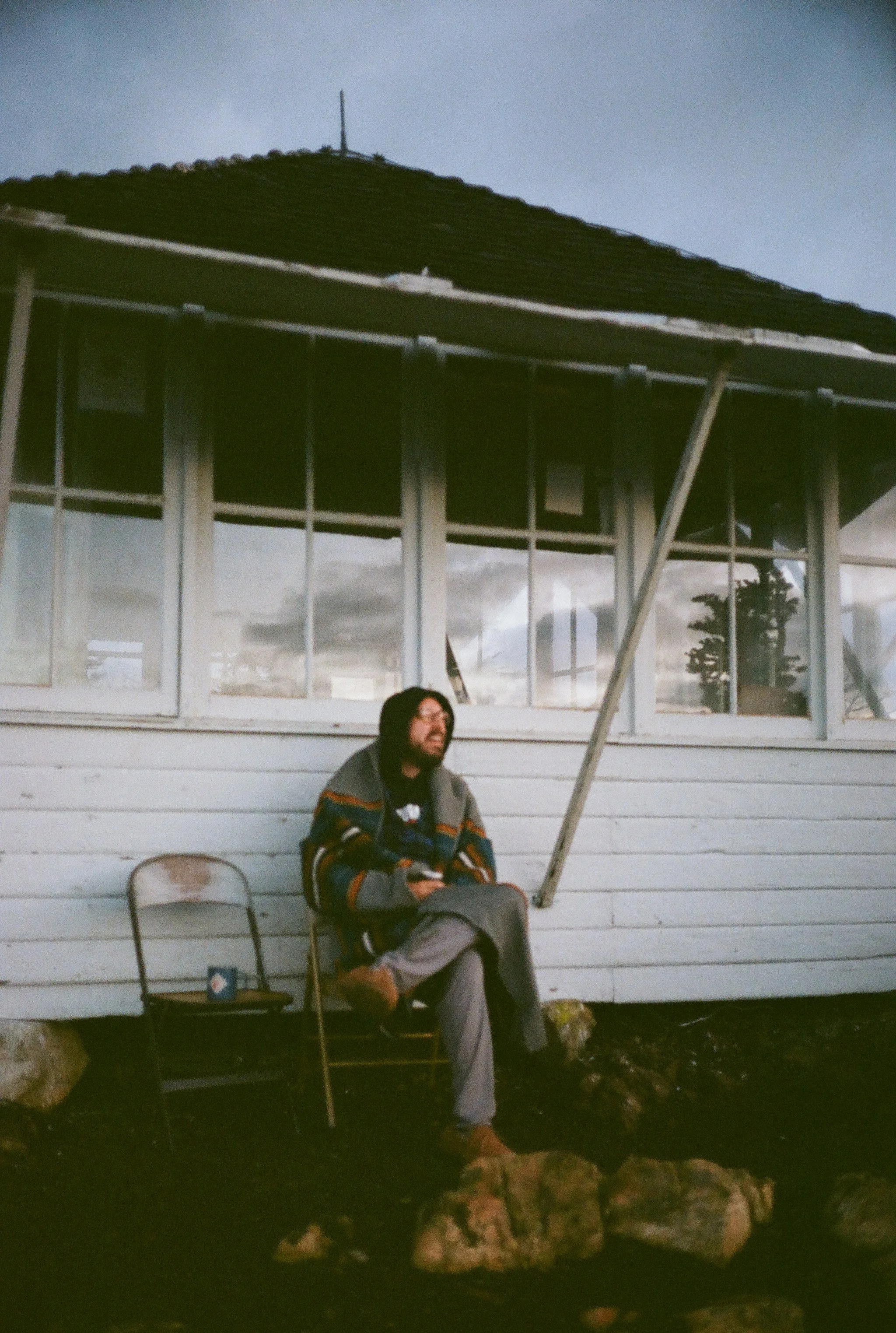 A man with long dark hair and glasses sitting on a foldable chair outside a white house with large windows. He is wearing a colorful striped sweater, gray pants, and brown shoes. There is a small table with a coffee cup next to him. The house has a r