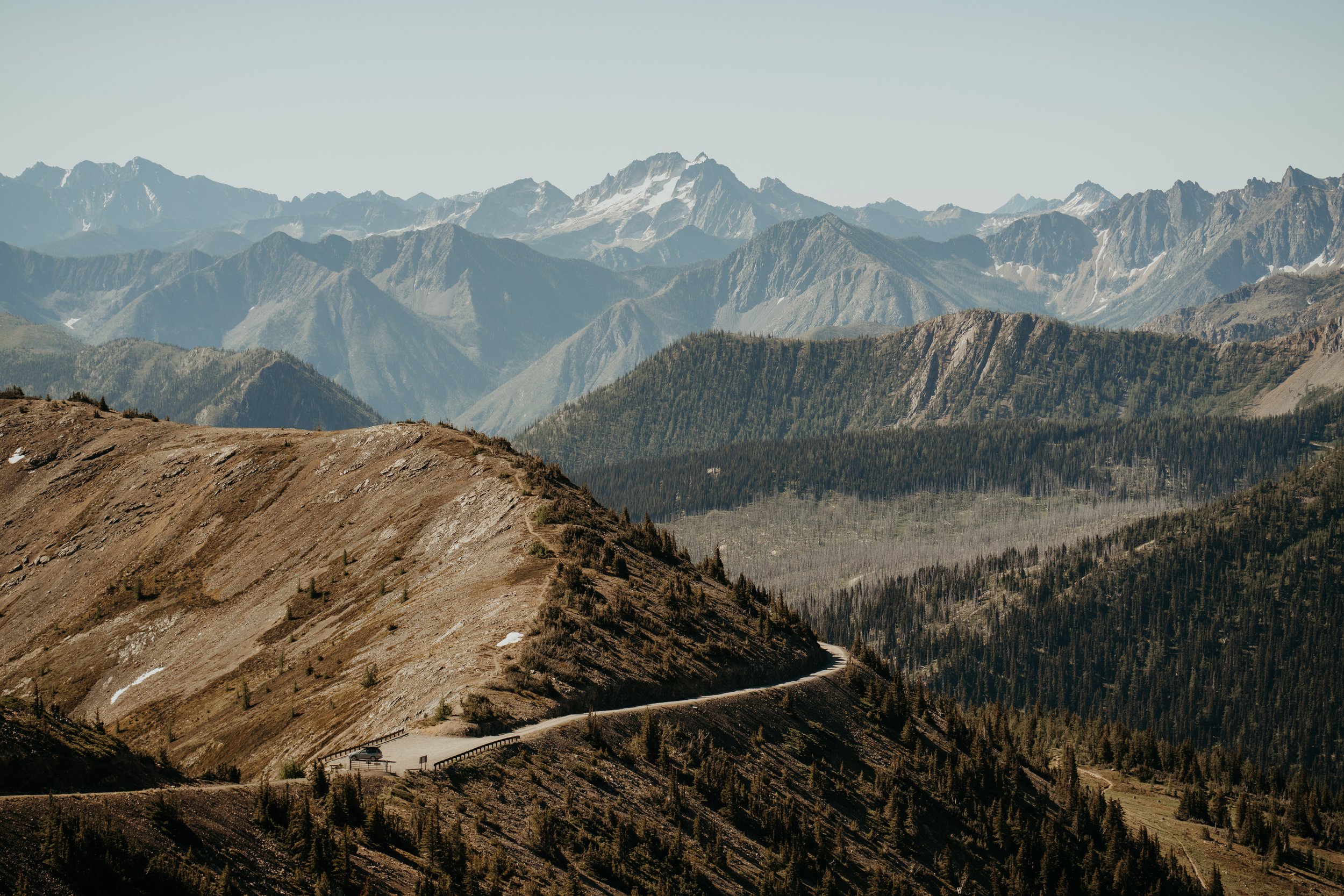 Mountain landscape with a winding road, forested slopes, and snow-capped peaks in the distance.