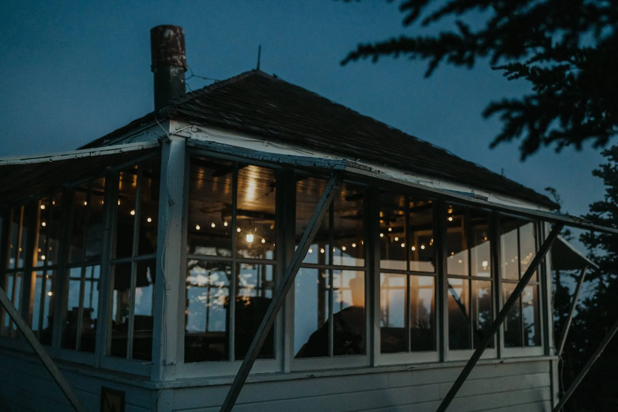 A cozy house with large windows illuminated from inside, seen at dusk with a dark blue sky and surrounding trees.