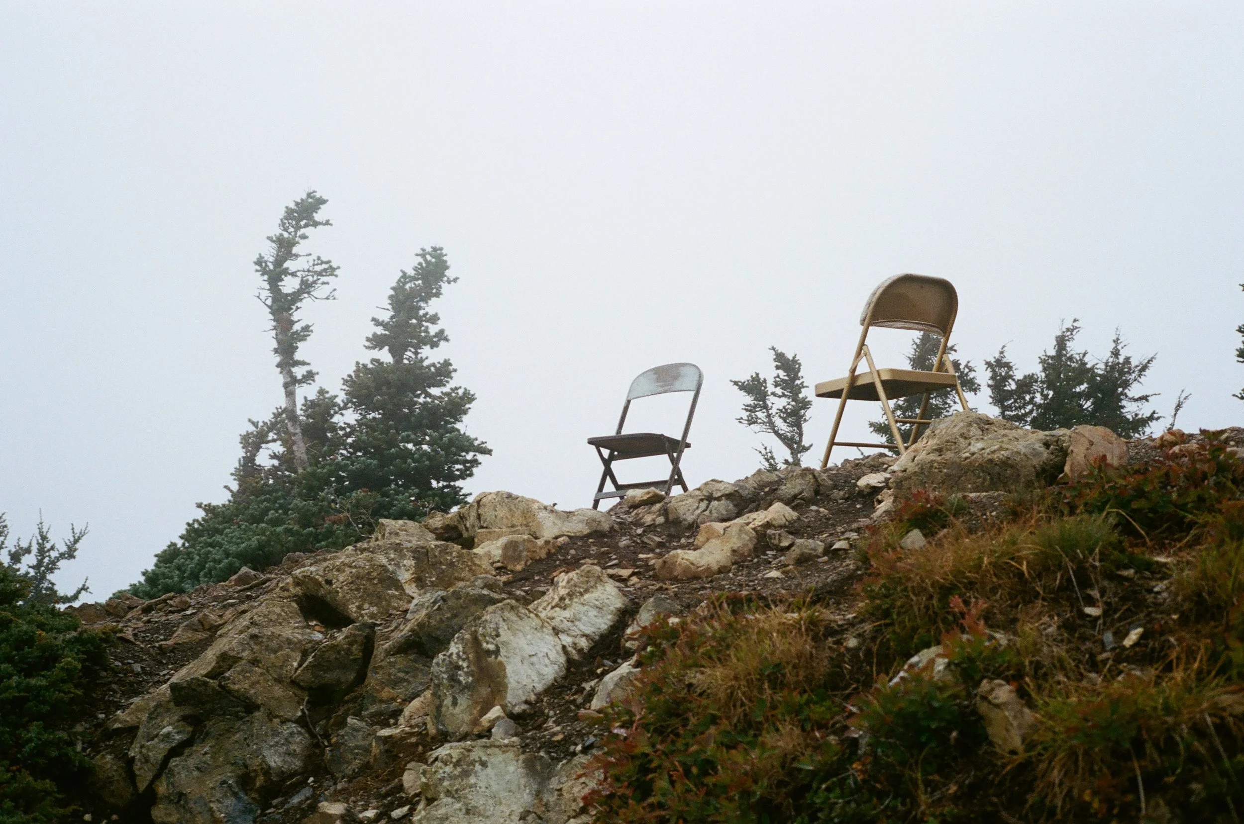 Two chairs on rocky terrain with evergreen trees in the background under foggy, overcast sky.