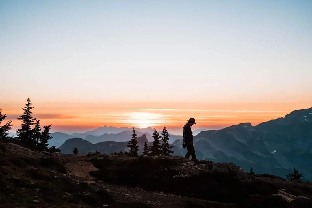 A hiker walking on a mountain trail during sunset, with pine trees and mountain ranges in the background.