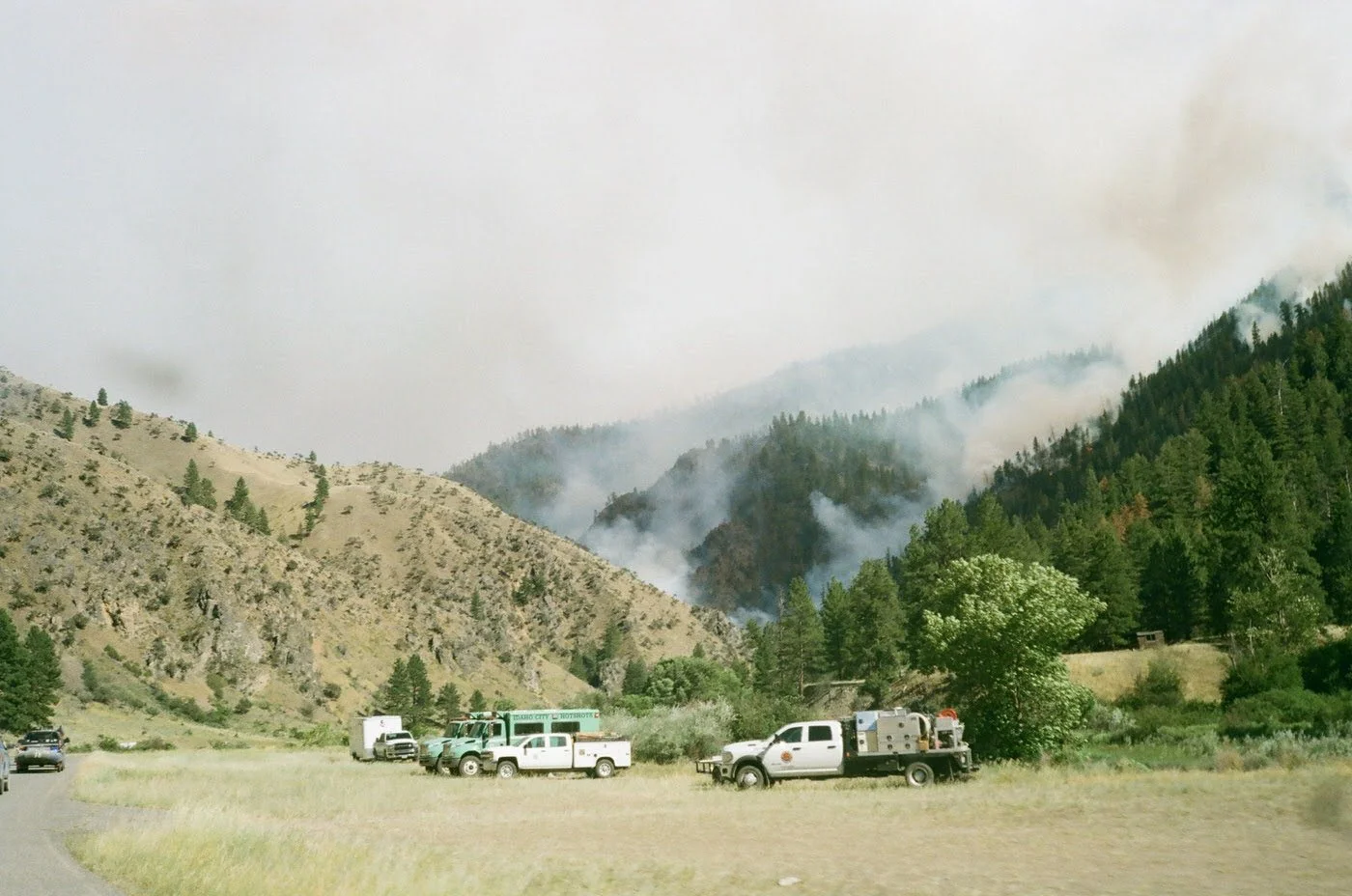Fire crews in trucks monitor a mountain wildfire with smoke rising from the forested hillside.