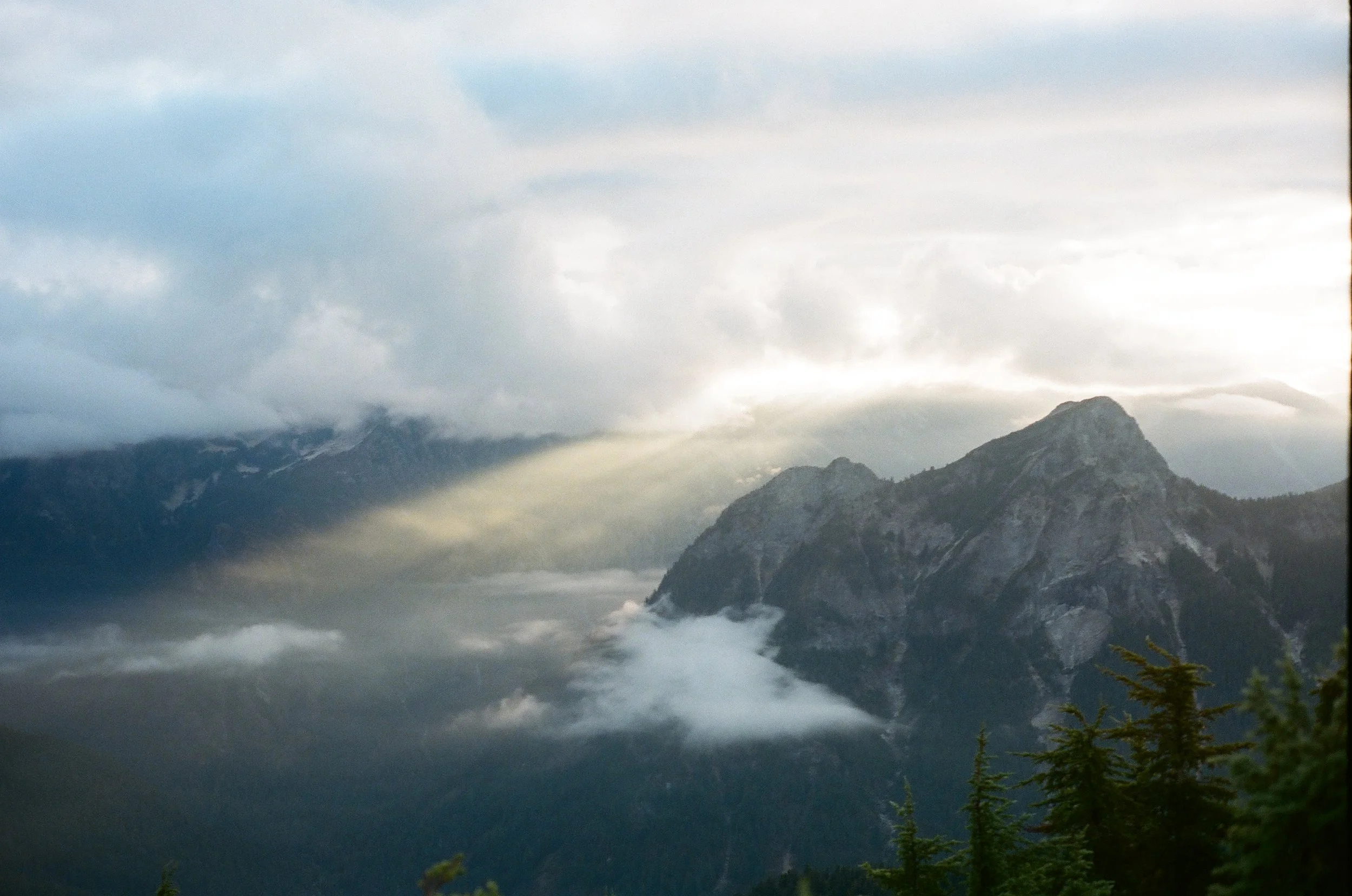 Mountain landscape with clouds and a sunbeam shining through the clouds over a forested mountain range.