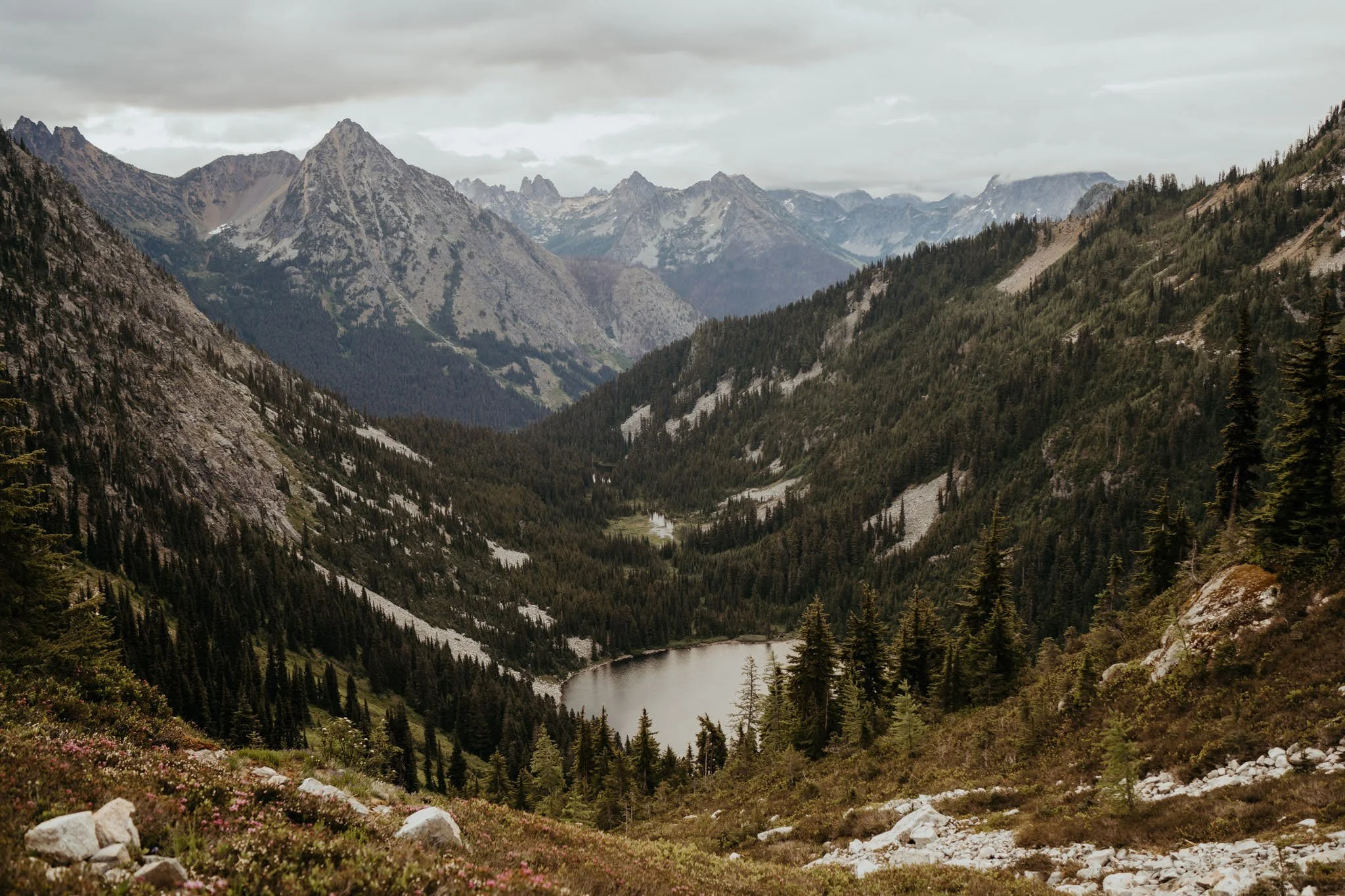 A mountainous landscape with a lake in a forested valley, surrounded by steep, rocky slopes and distant snow-capped peaks under cloudy skies.