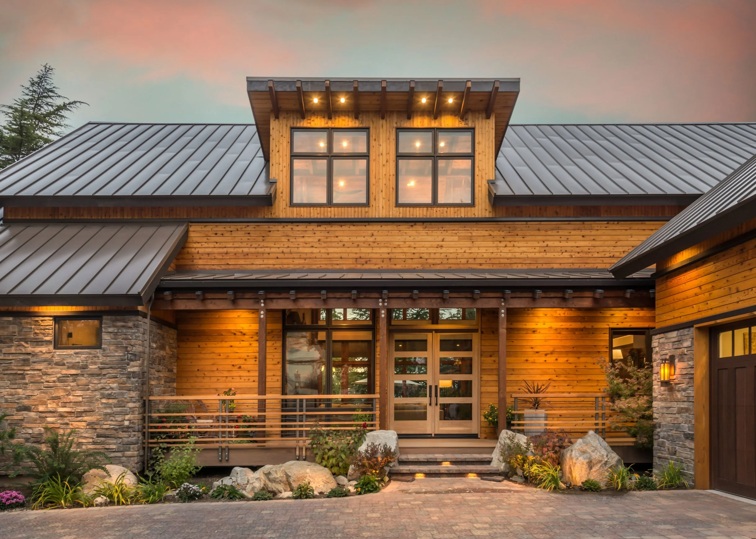 A modern house with wooden siding, stone accents, and a metal roof, illuminated by warm outdoor lighting at dusk. The house has a small front porch with plants and large rocks, and a parking area with paving stones.