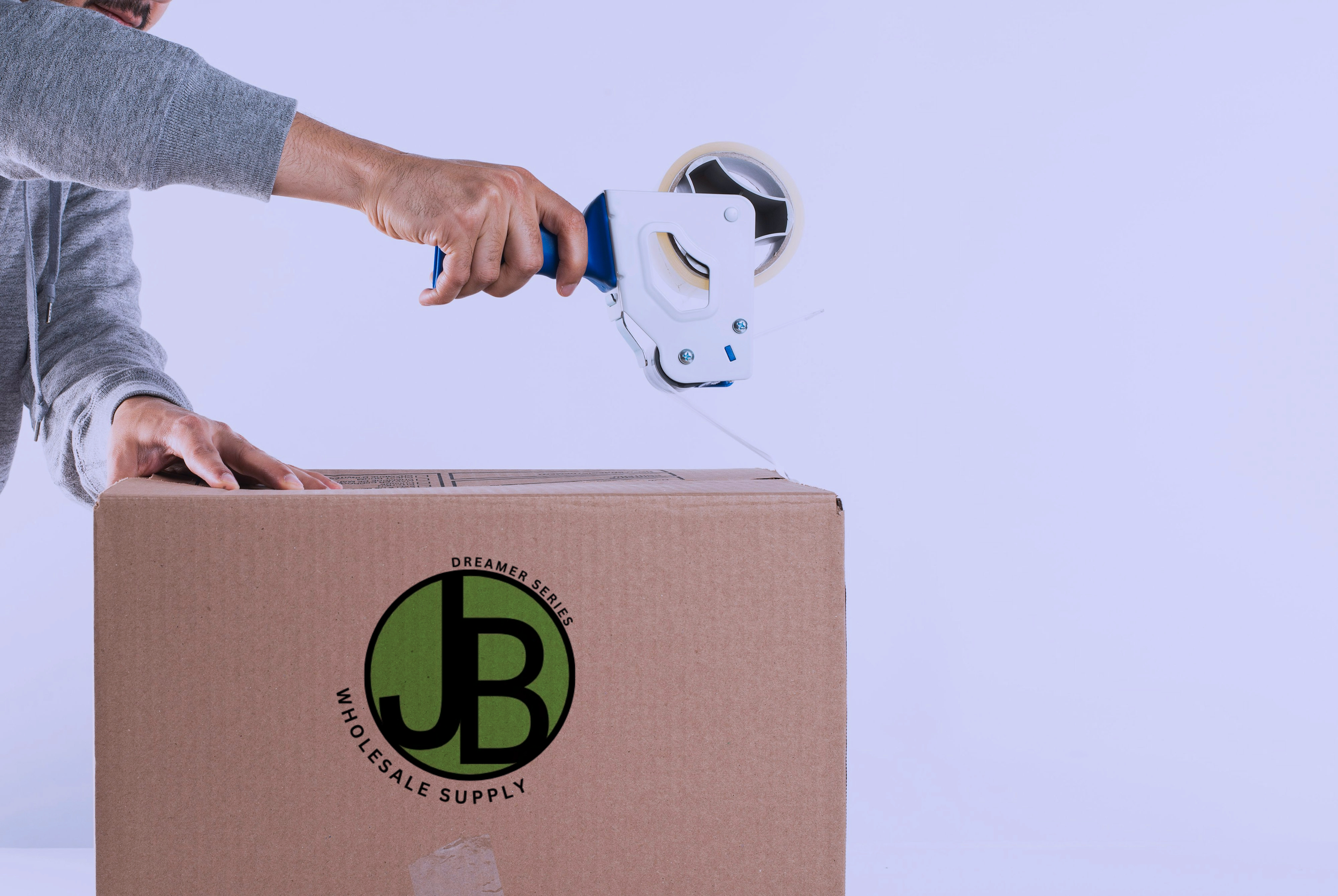 A person in a gray hoodie is using a tape dispenser to seal a cardboard box. The box has a logo with a green circle and black lettering.