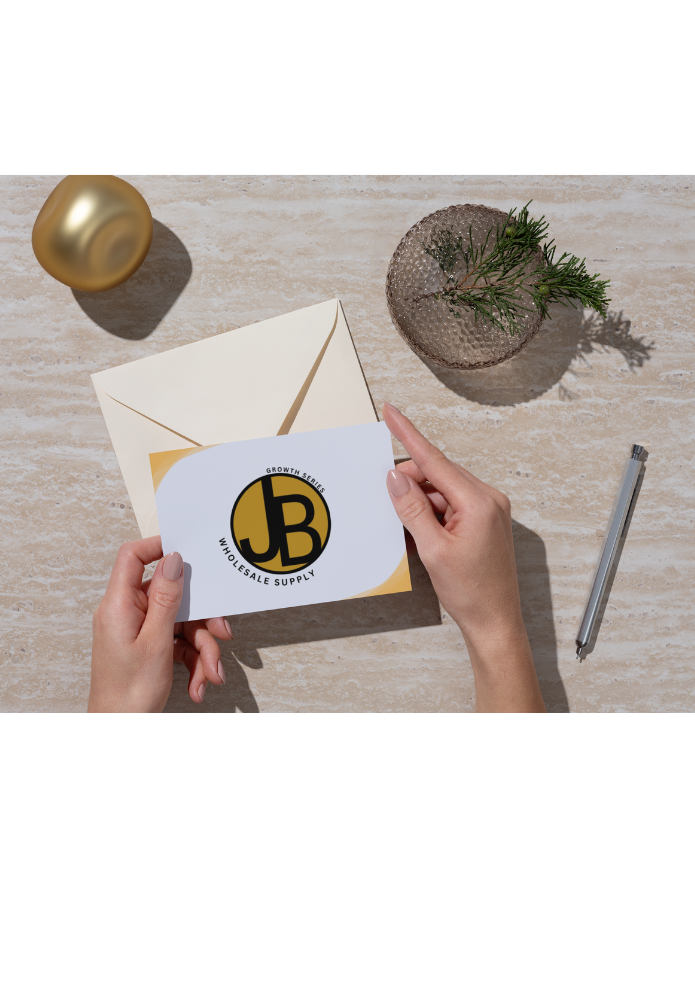 Person holding an envelope and a card with a logo that says 'JB Wholesale Supply'. The desk has a gold decorative item, a small round glass vase with greenery, and a pen.