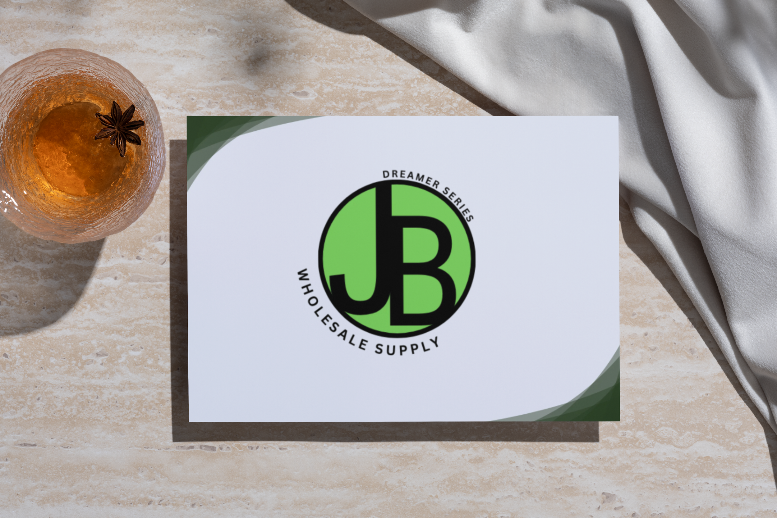 A glass of amber-colored beverage with a star anise on top, next to a white sheet of paper with a green and black logo that reads 'Dreamer Series Wholesale Supply' on a light-colored textured surface.
