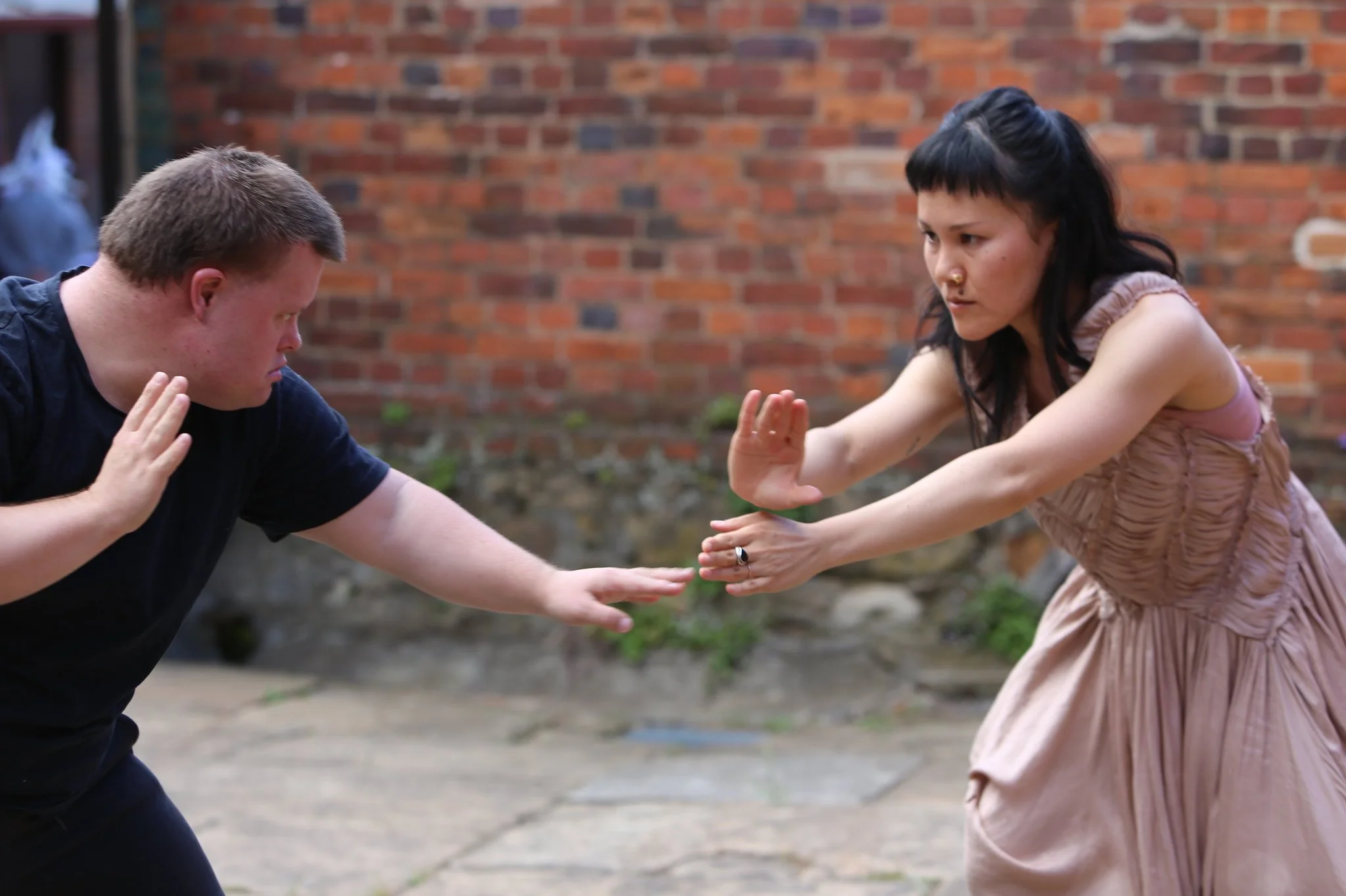 Two people with arms outstretched in front moving dance-like, martial arts-like. They are outdoors in front of a brick wall.