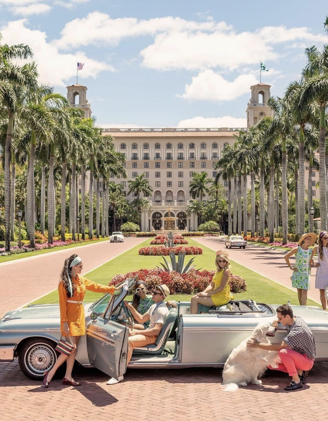 Group of women and a man with a dog in front of the Biltmore Hotel in Miami, Florida, with tall palms, a fountain, and a grand hotel building in the background.