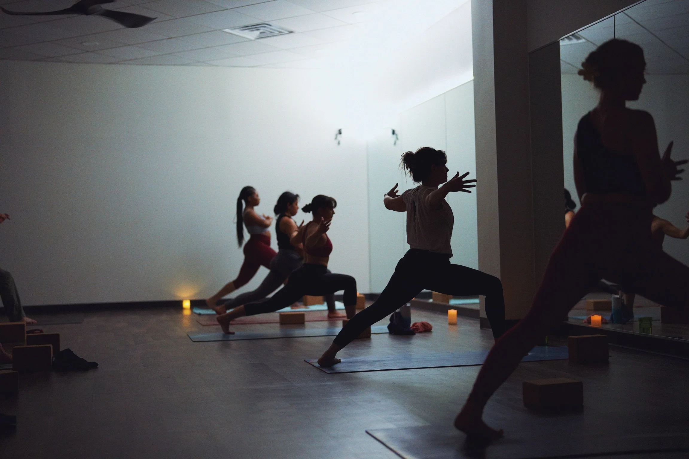 Group of people practicing yoga in a dimly lit studio with candles, facing a mirror, in a Warrior stance with arms extended.