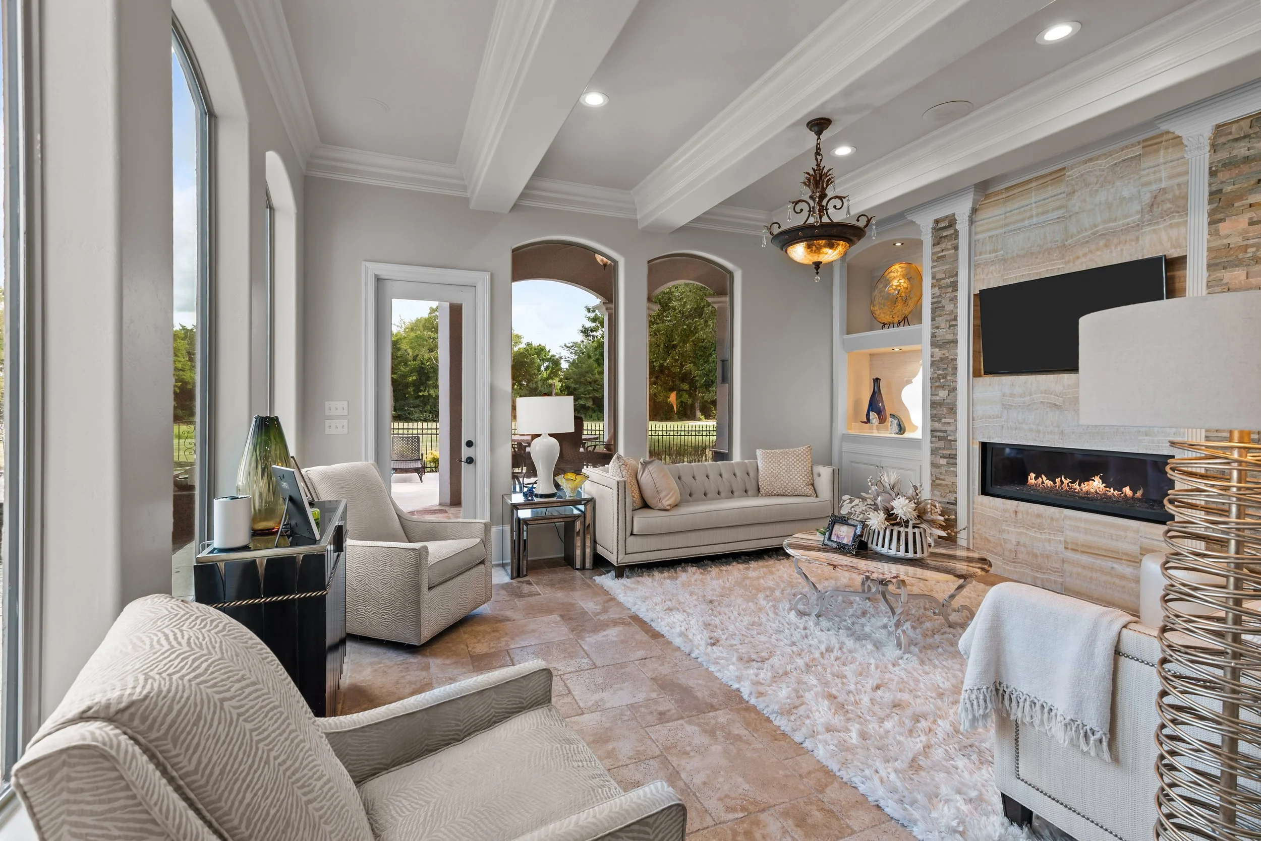 Living room with large windows, beige furniture, a fireplace, flat-screen TV, chandelier, and outdoor view.