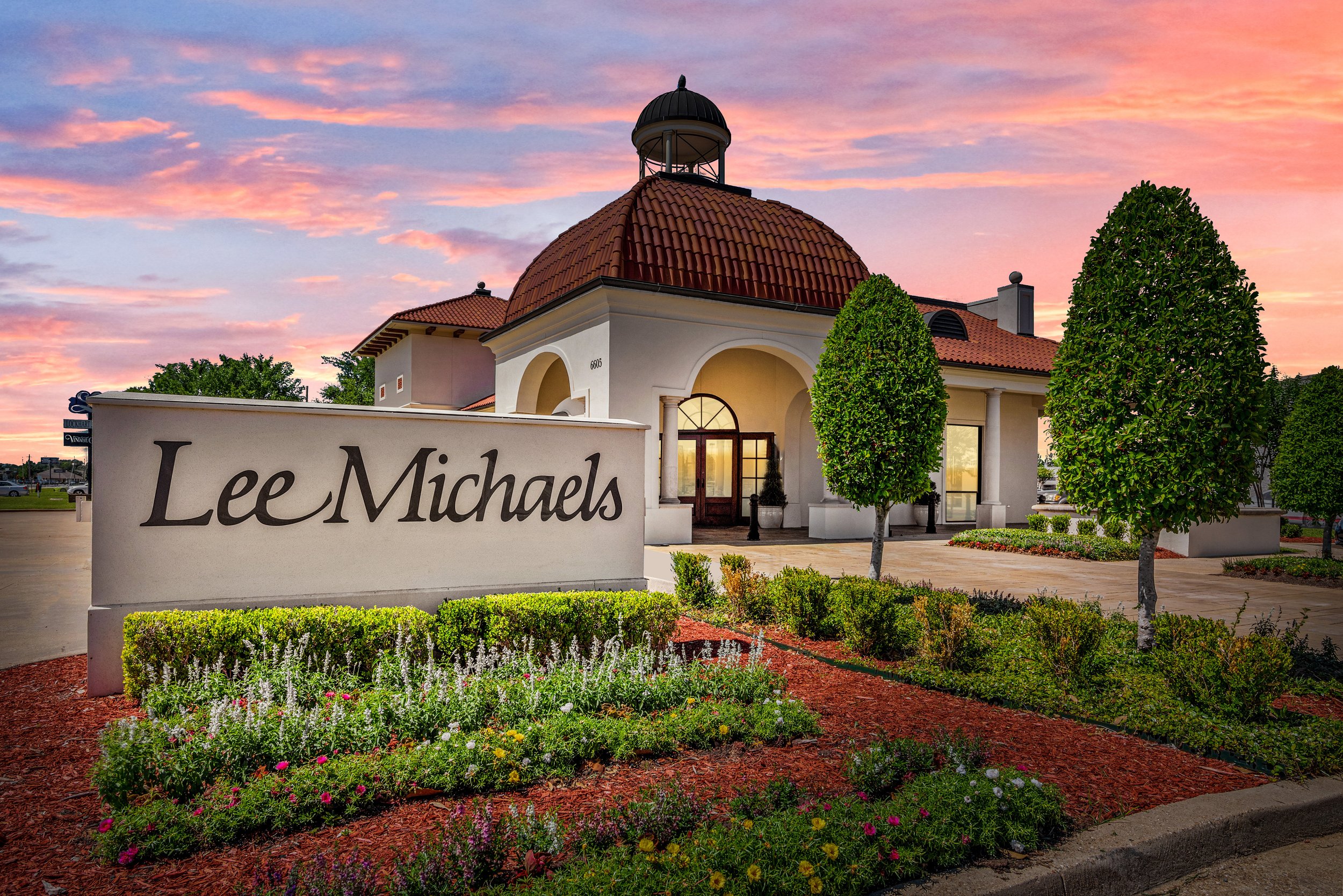 The front of Lee Michaels store with a sign, surrounded by landscaped flower beds and two tall, sculpted trees, under a colorful sunset sky.