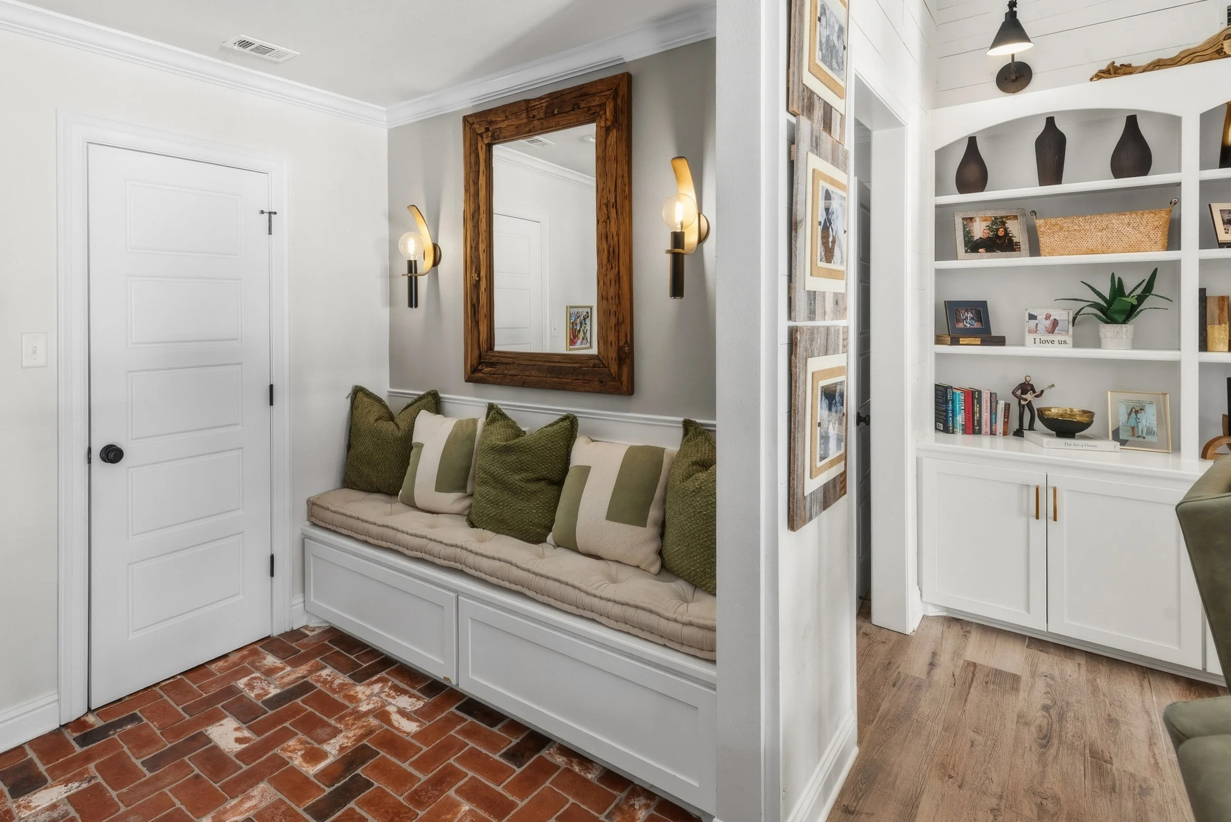A cozy living area with a built-in white bench under a large wooden-framed mirror, with green and beige pillows. To the right, there are white shelves with various decorative items, framed photos, and books, and a partial view of a green chair nearby