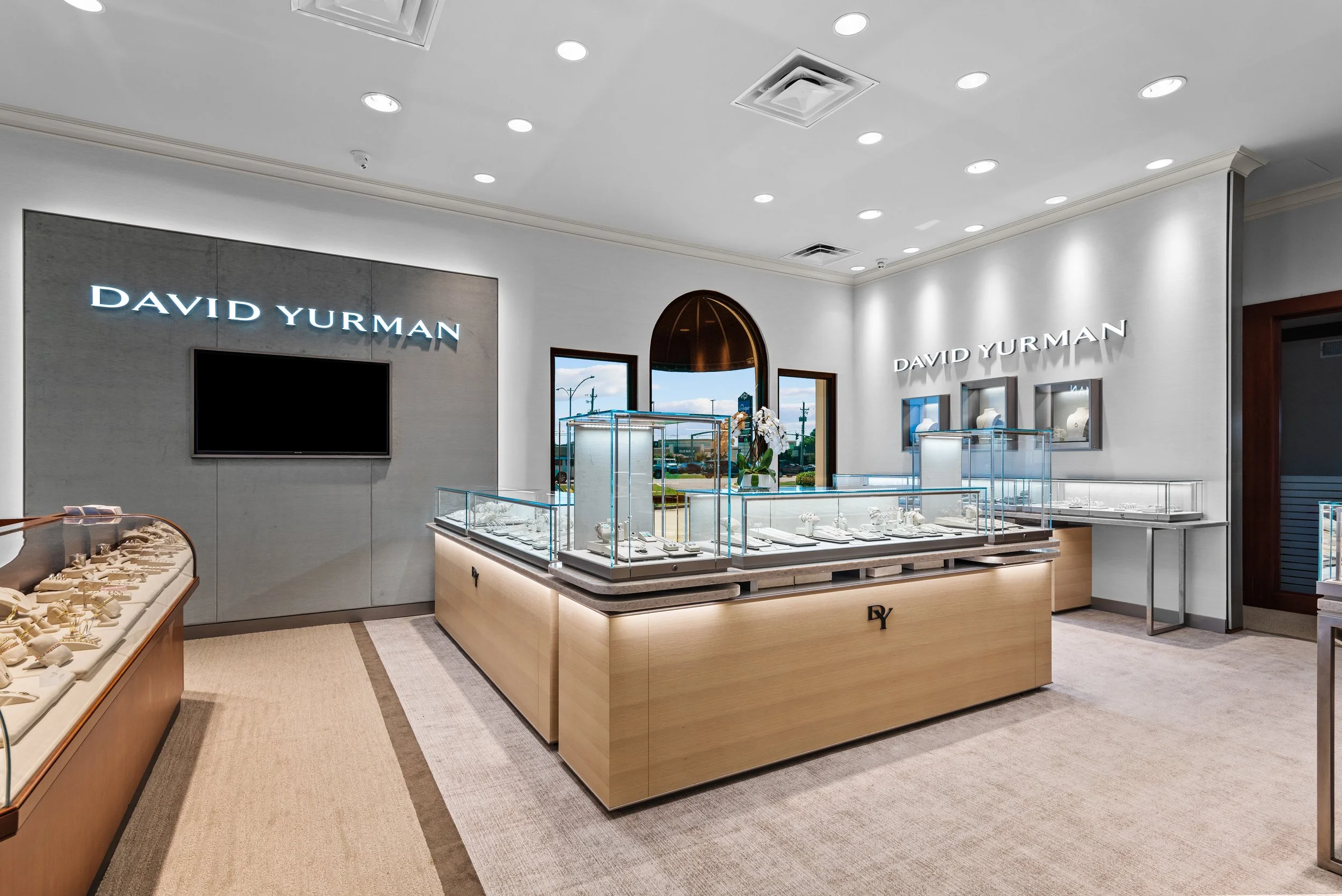 Inside a jewelry store with display cases containing jewelry, a large wall sign reads 'David Yurman,' with windows showing a street view outside.