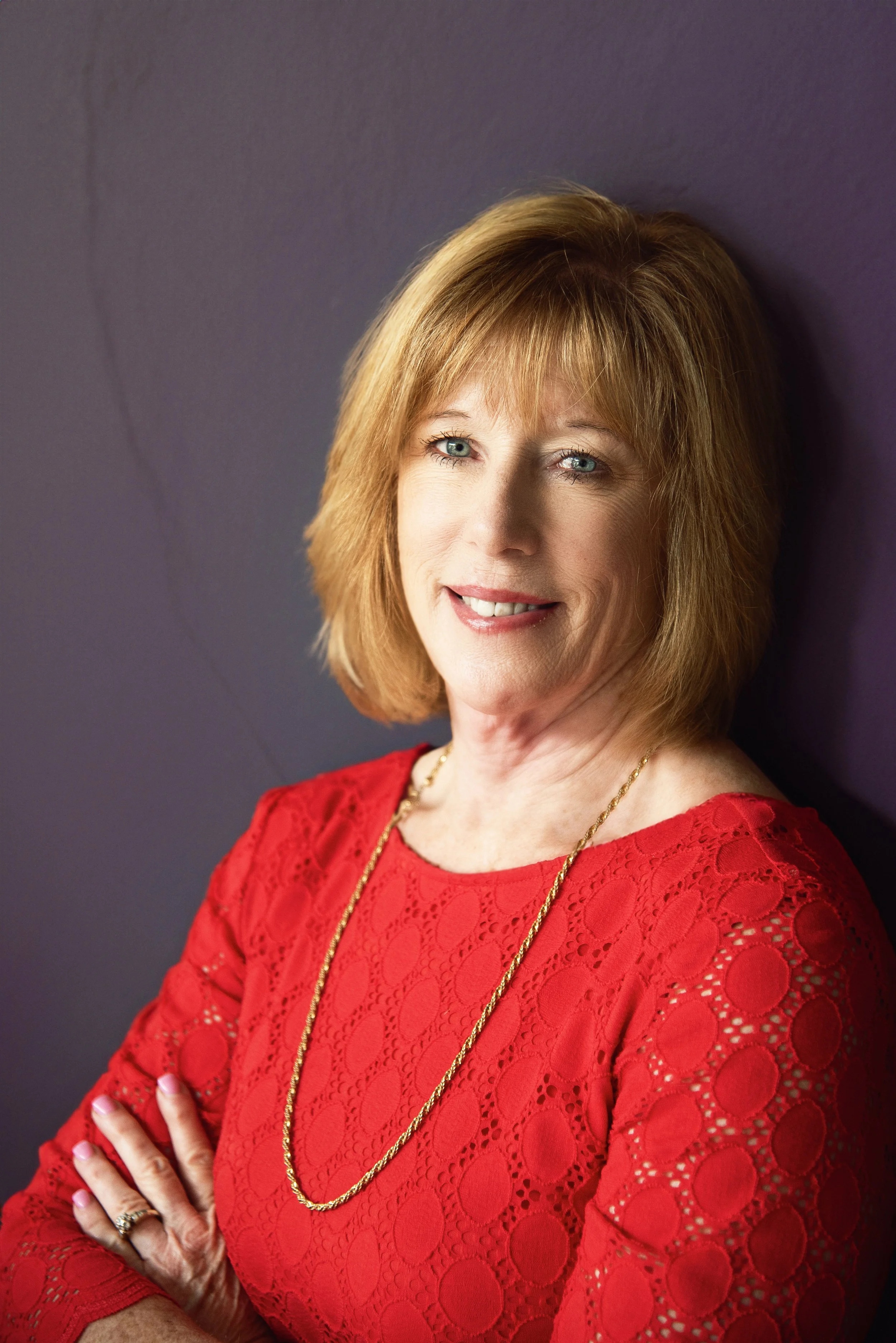 A woman with red hair wearing a red lace dress, a gold necklace, and rings, standing against a purple wall, smiling and crossing her arms.