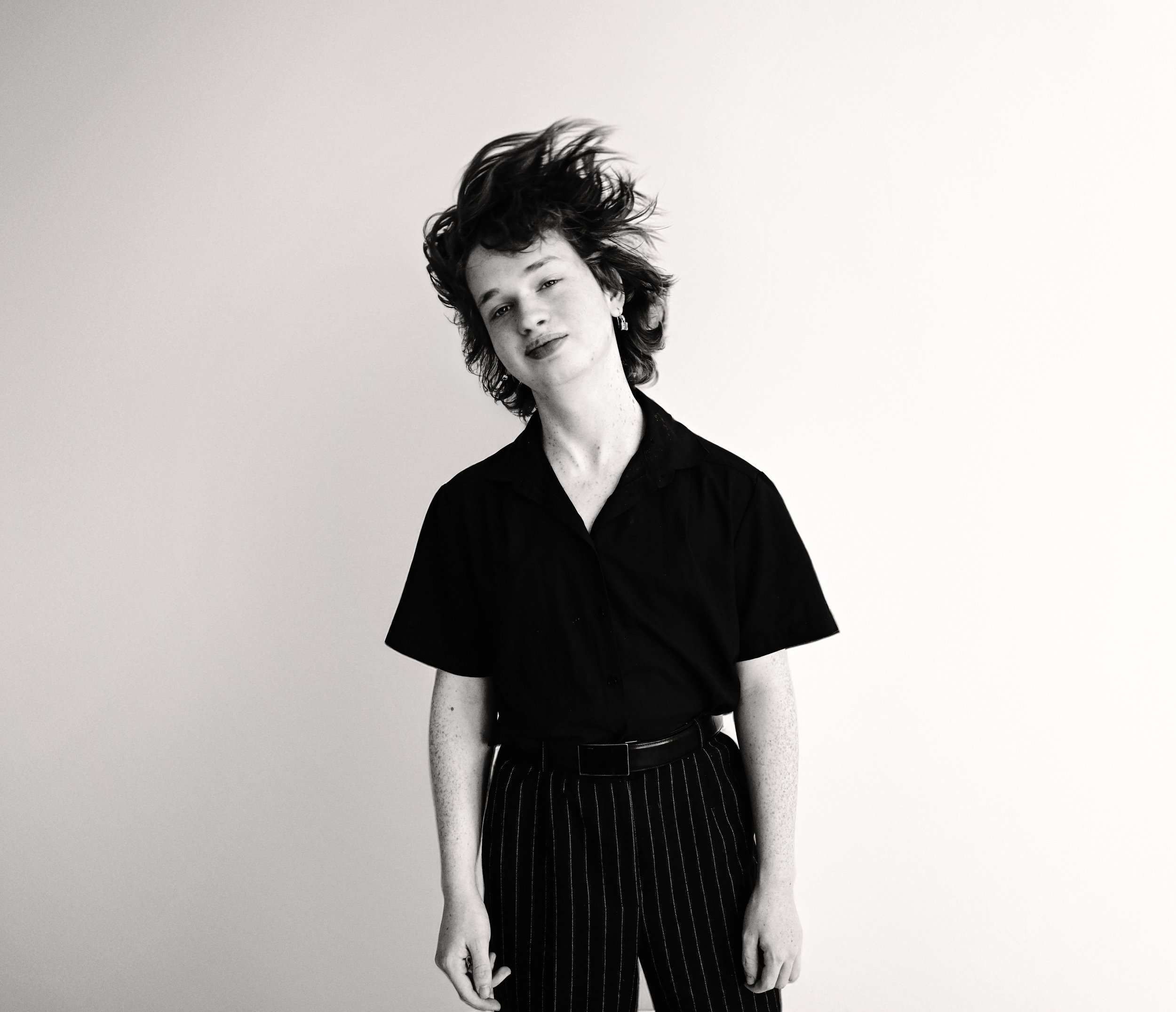 Black and white photo of a person with short, curly hair, wearing a black shirt, standing against a plain white background. The person is looking at the camera with a slight smile.