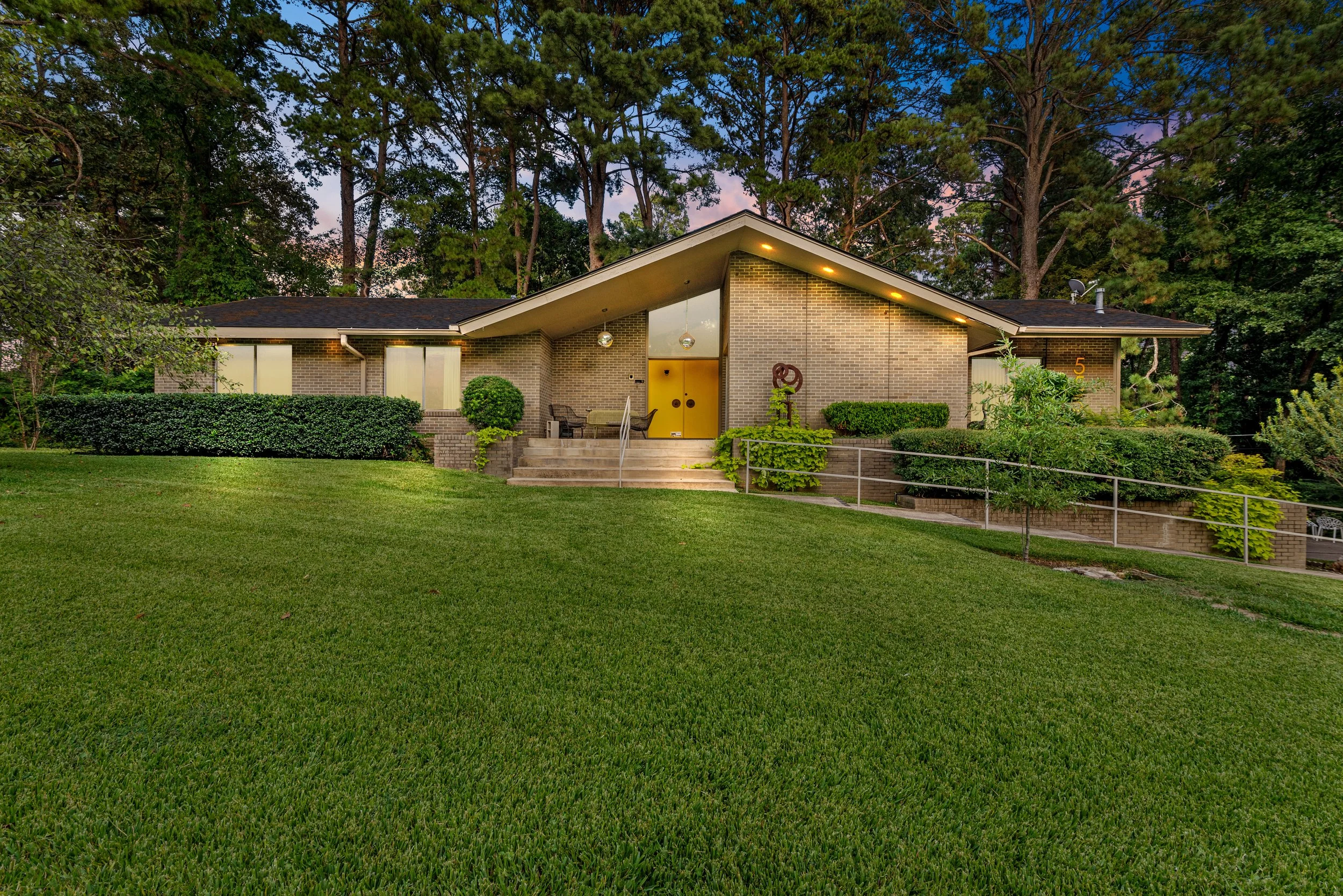 A mid-century modern brick house with large windows, a yellow front door, and outdoor seating, surrounded by well-maintained lawn and lush trees at dusk.