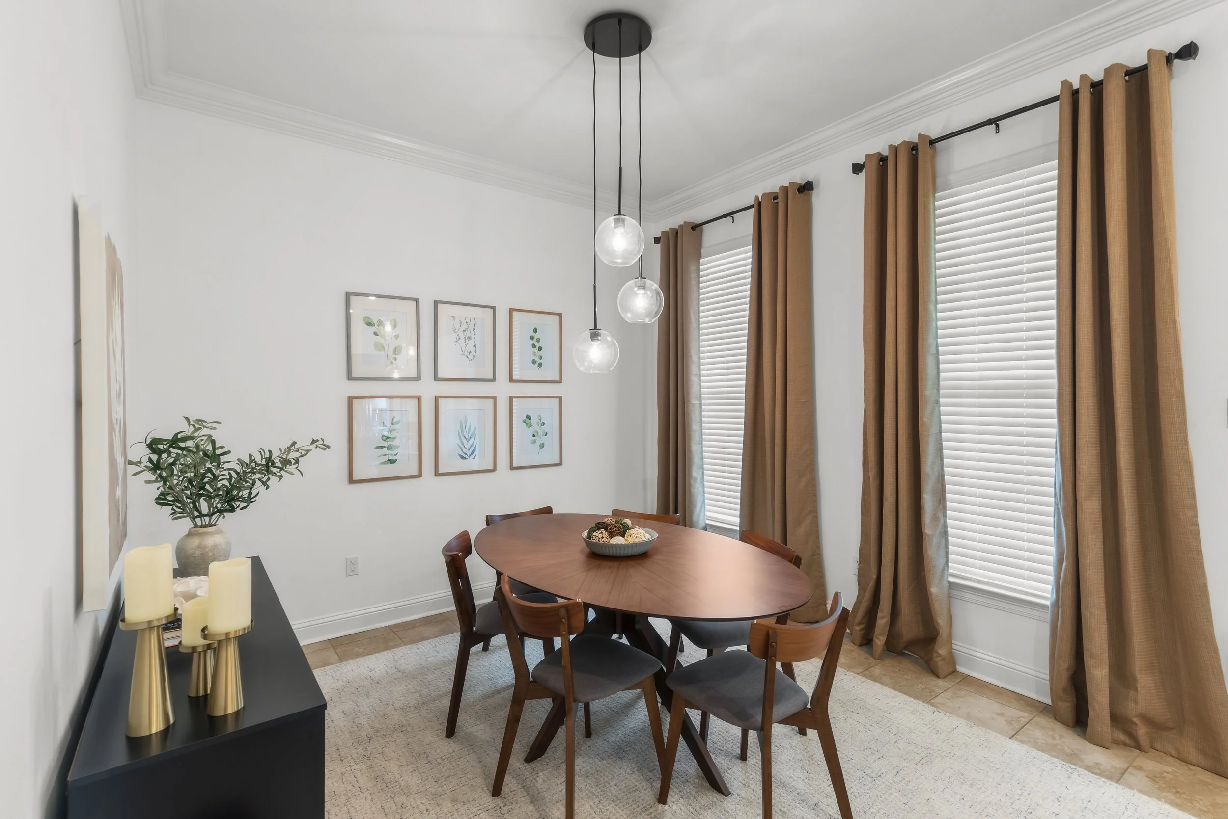 A dining room with a round wooden table, six chairs, and a wall decorated with six framed botanical prints. There are two large windows with beige curtains, and a modern chandelier with three globes hanging above the table. A black sideboard holds ca