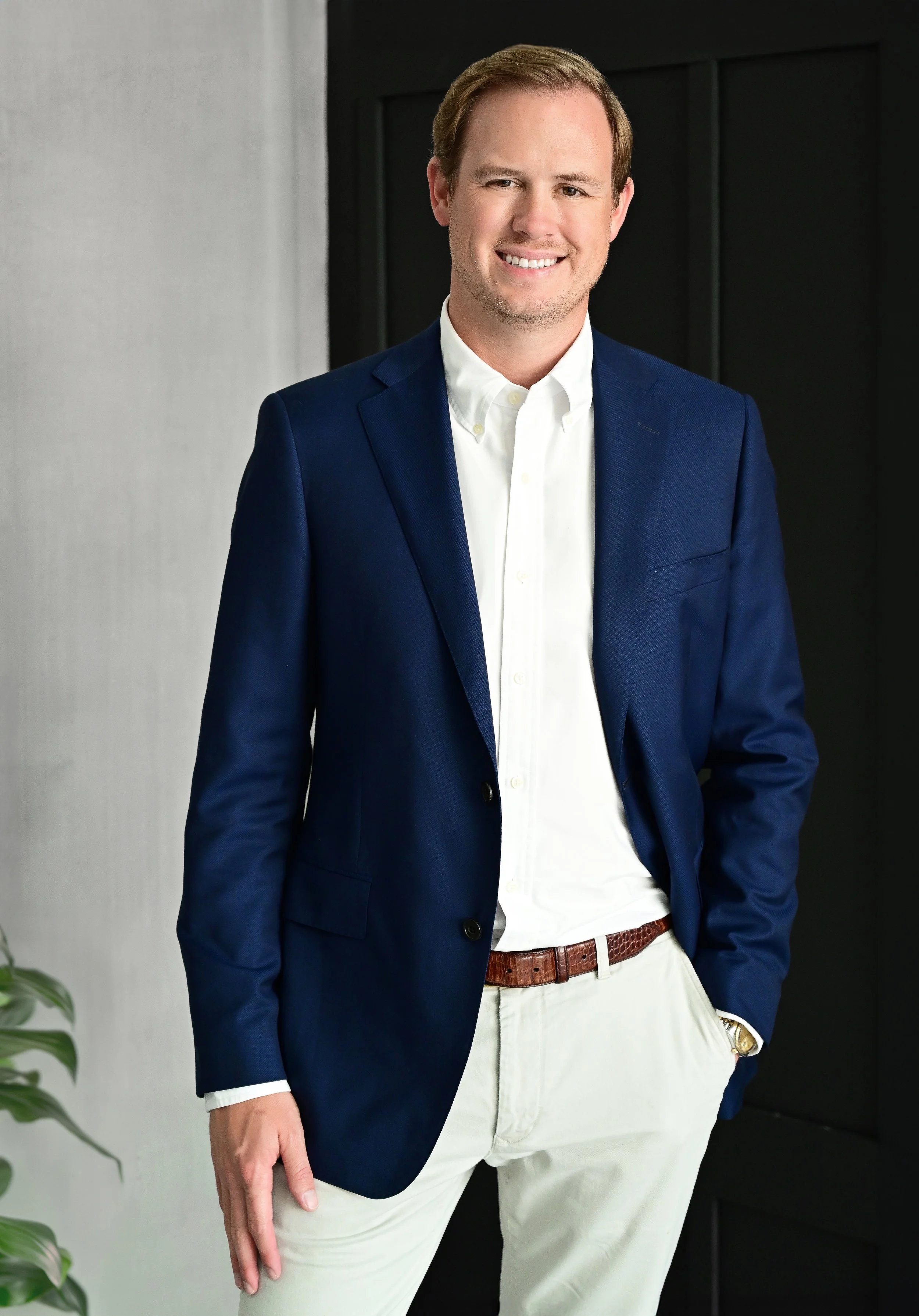 A man in a blue blazer, white shirt, and beige pants standing indoors with a dark background and a plant visible on the lower left.