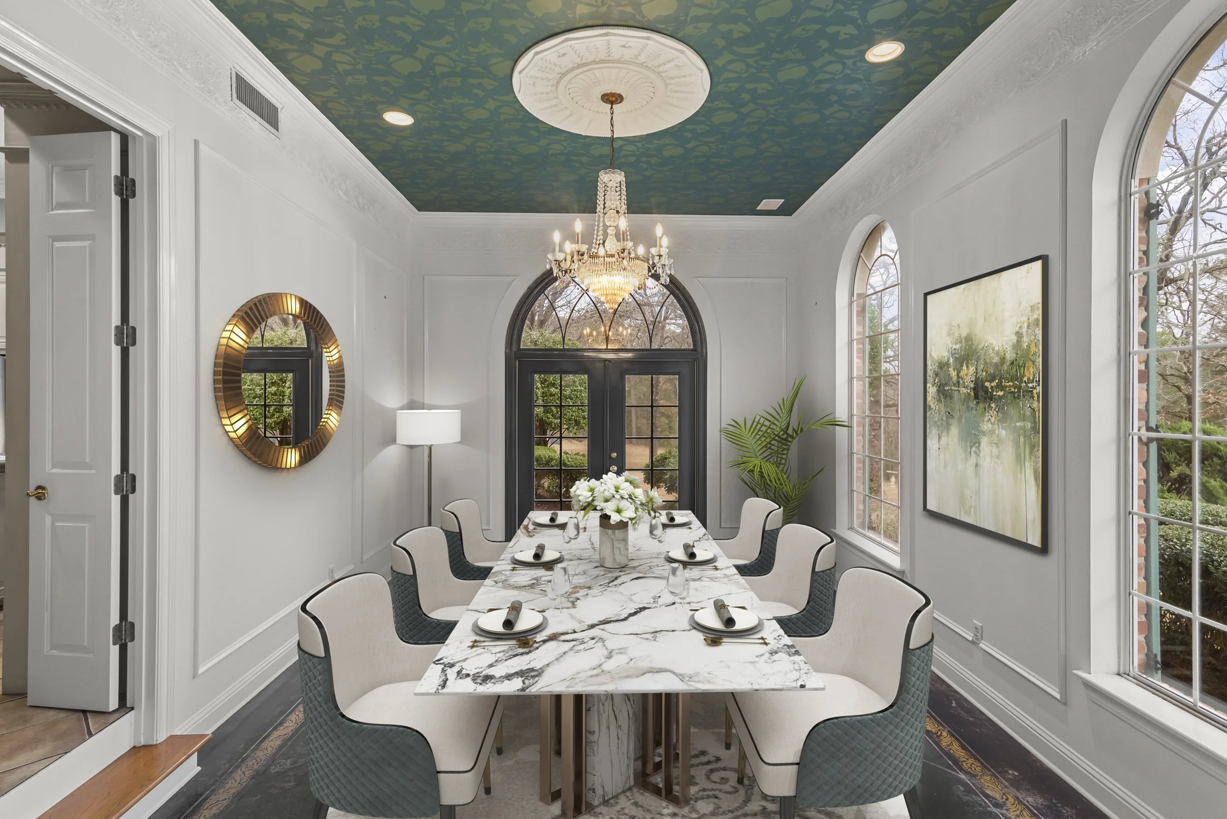 A dining room with a long marble table set for eight people, surrounded by modern white chairs. The room features large windows, a chandelier hanging from a decorative ceiling, a round mirror, a potted plant, and artwork on the walls. A bouquet of fl