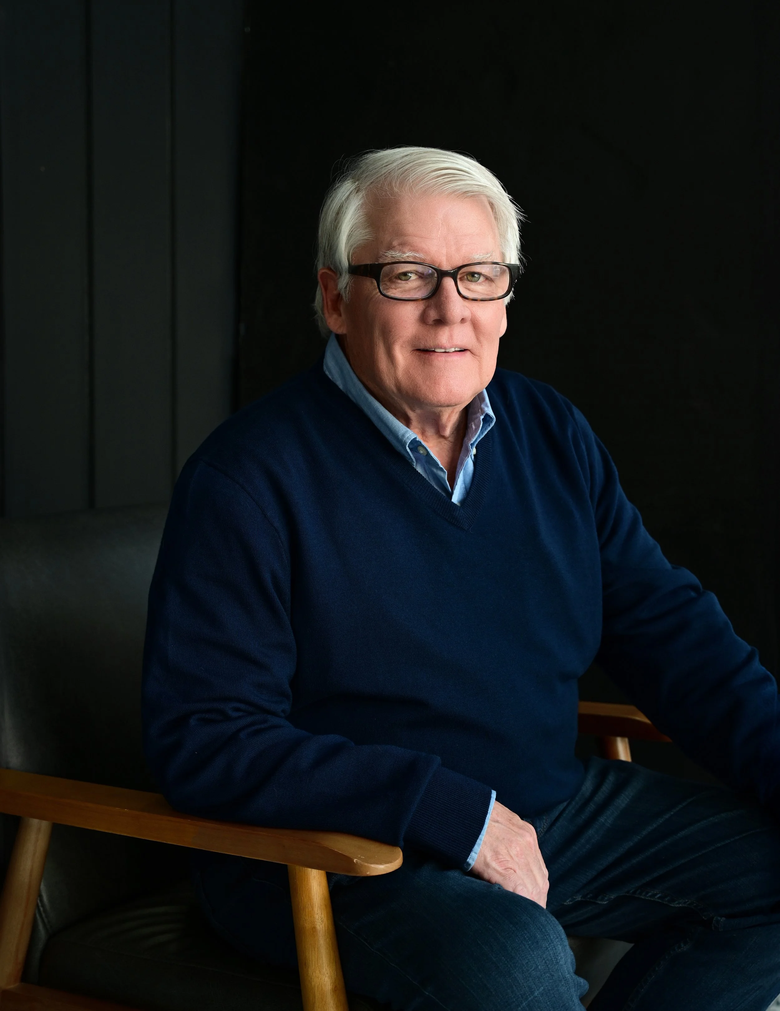 An elderly man with gray hair and glasses sitting on a wooden chair, wearing a blue sweater over a collared shirt, against a dark background.