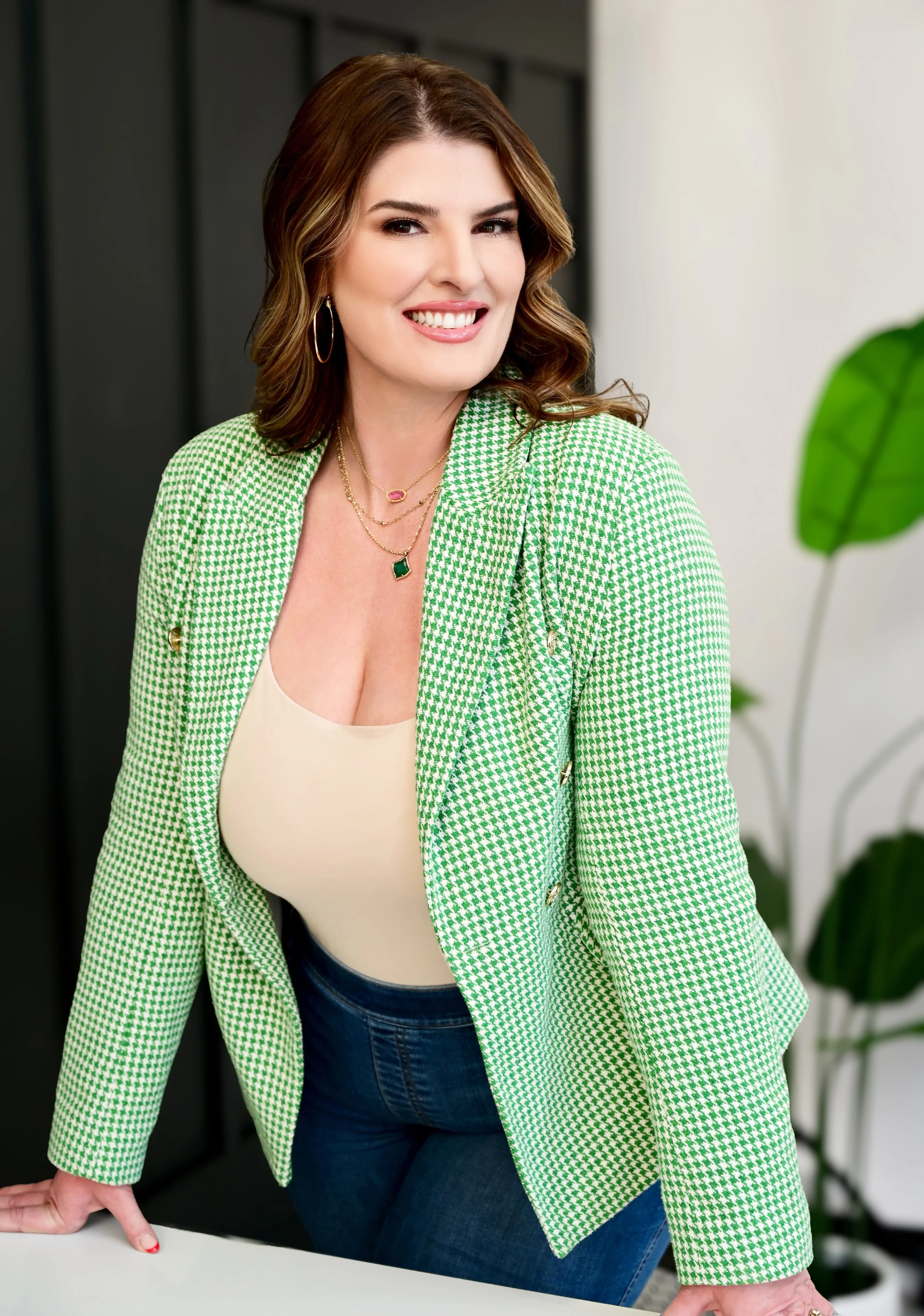 A woman with brown hair wearing a green and white houndstooth blazer, cream top, and blue jeans, smiling and leaning on a white surface with one hand, in an office setting with plants in the background.
