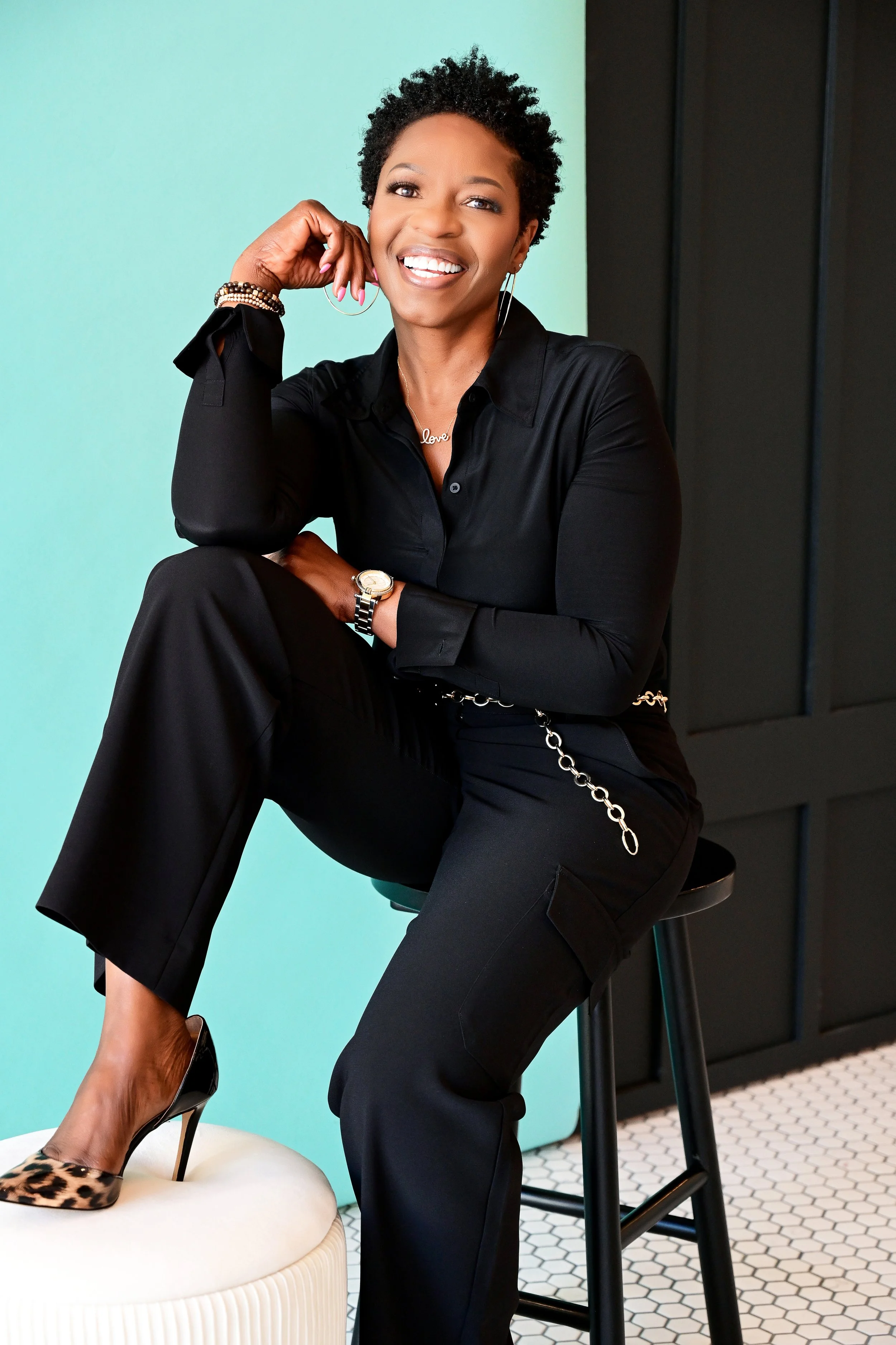 A woman sitting on a black stool, smiling, wearing a black blouse, jewelry, and leopard print high heels, with a mint green wall in the background and black cabinets.