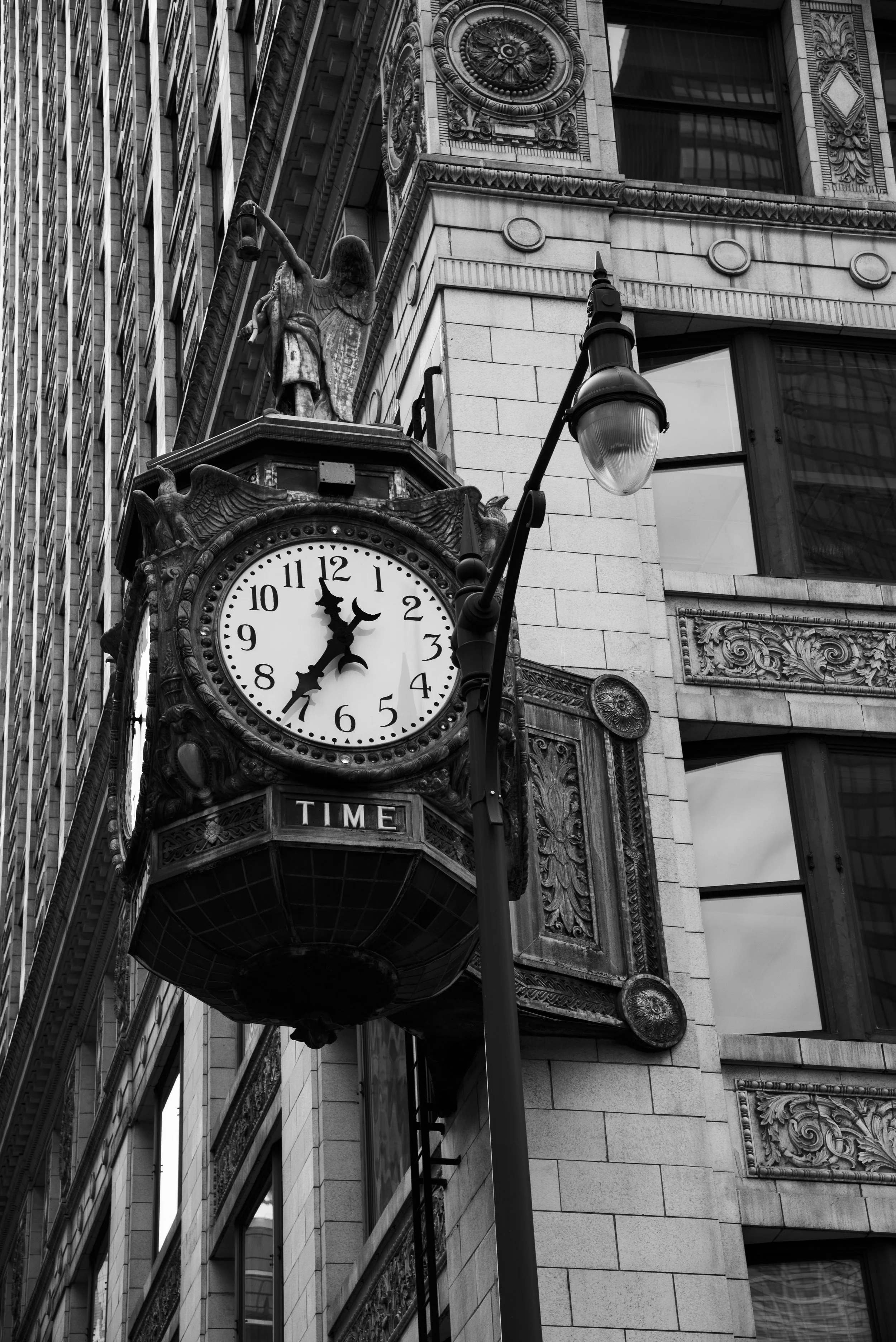 Black and white photo of a vintage street clock mounted on a building facade in Chicago, with ornate decorative elements, a street lamp, and tall office buildings with window reflections in the background.