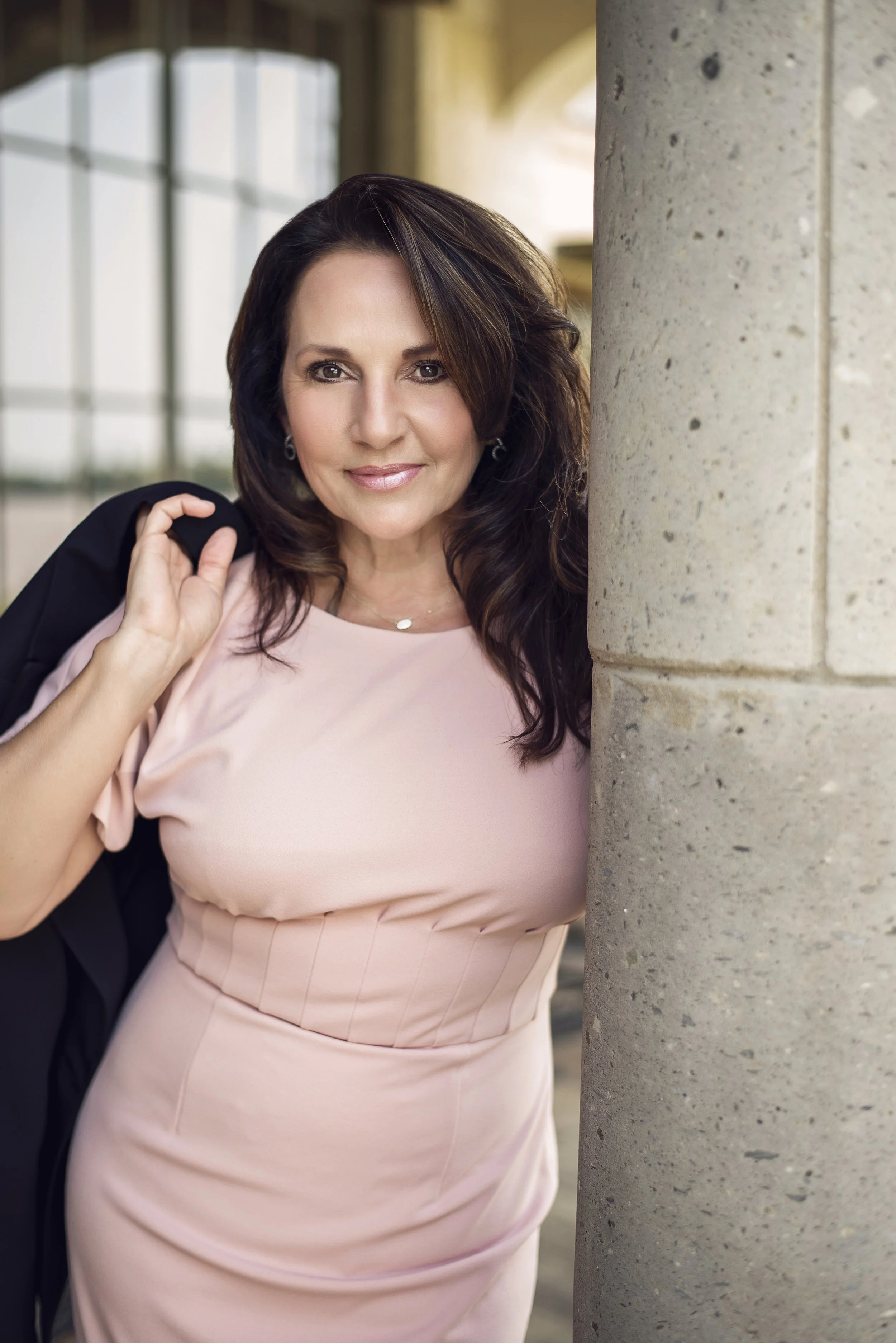 A woman with dark brown hair and a pink dress standing partially behind a stone pillar, looking at the camera with a slight smile.