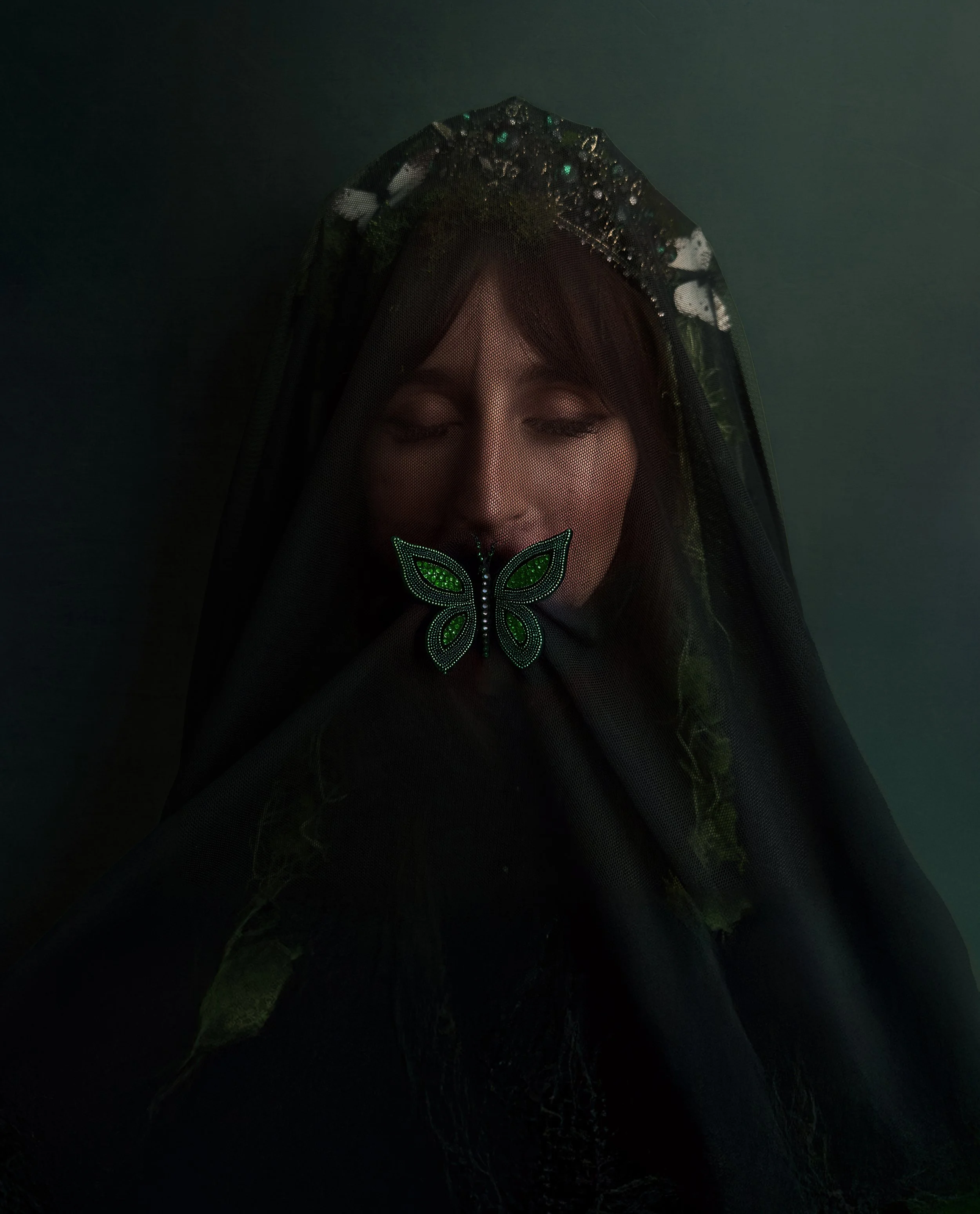 A person with closed eyes wearing a decorative butterfly mask, shrouded in black lace veil, with a dark background.