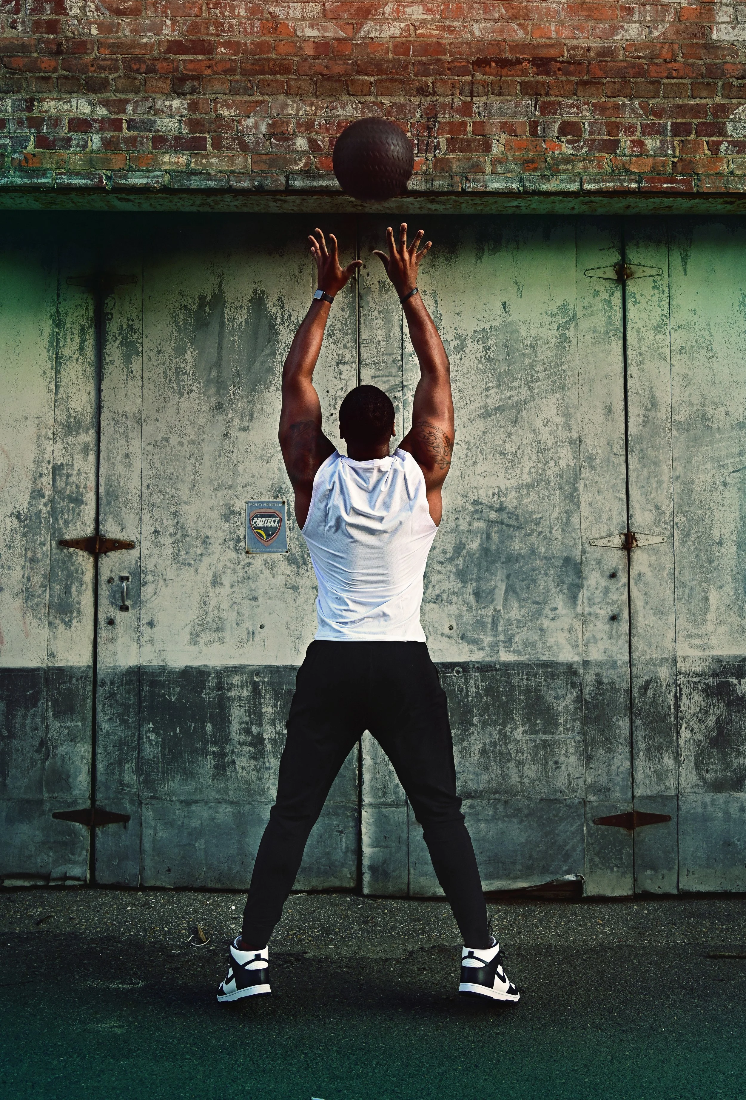 A man in athletic clothing practicing basketball shooting against a weathered green wall with a brick upper section.