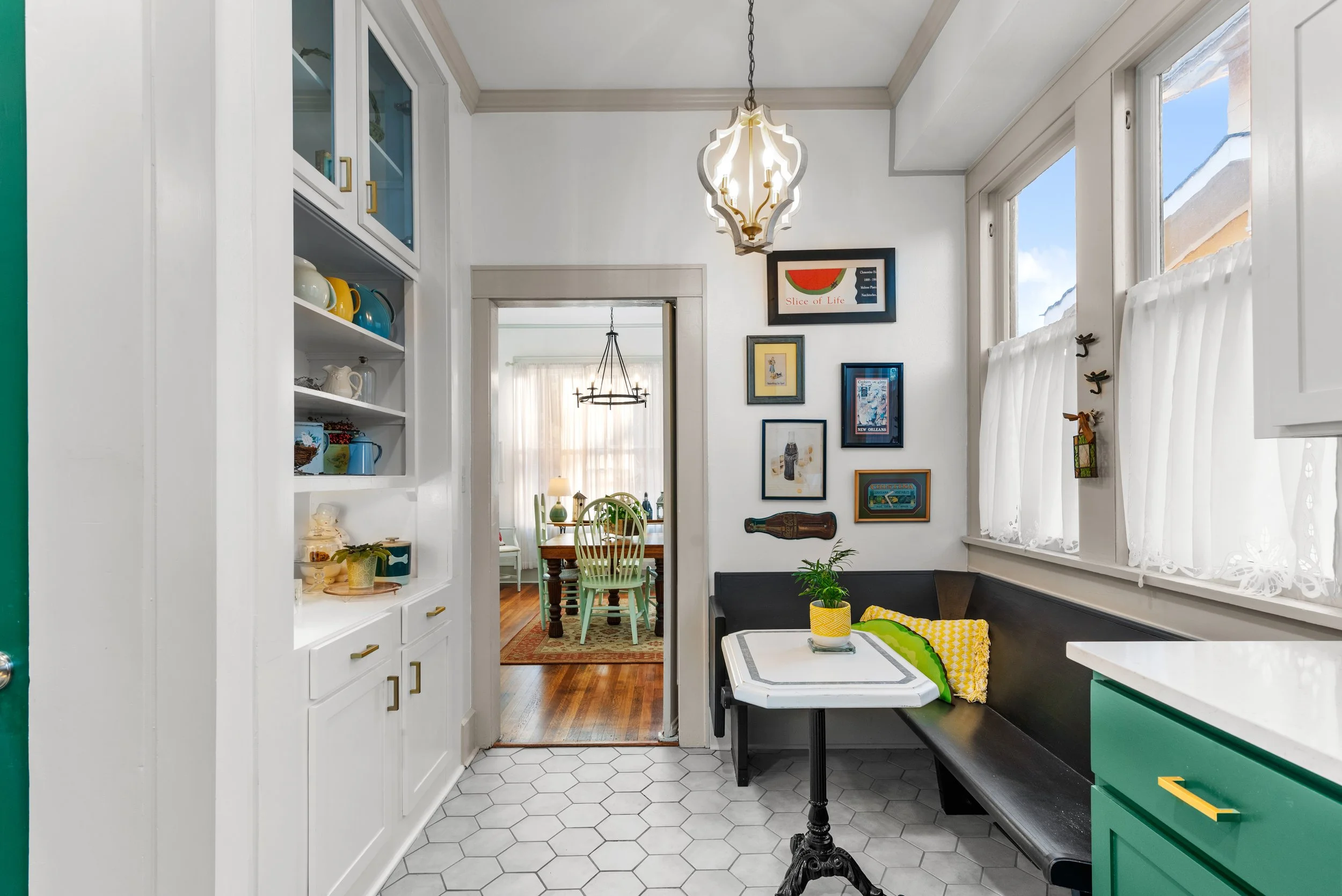 A cozy kitchen with white cabinets, hexagonal tile flooring, and a black bench seat. The walls are decorated with framed artwork, and a view into a dining area with a table and light-colored chairs is visible through an open doorway. A hanging light 