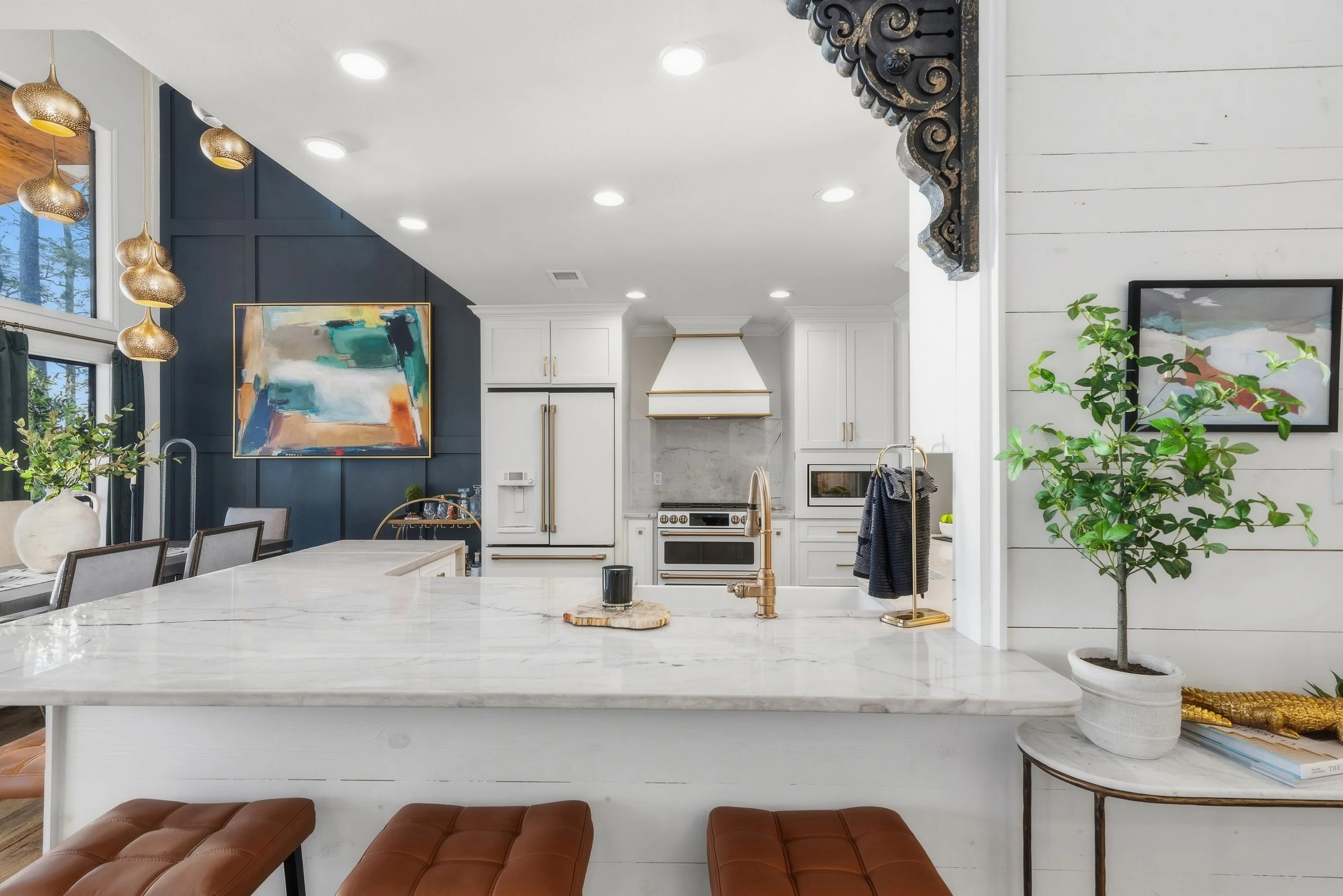 Modern kitchen with white cabinets, a marble island, gold hardware, and contemporary artwork on a navy blue accent wall. Includes pendant lights, potted plants, and windows with natural light.