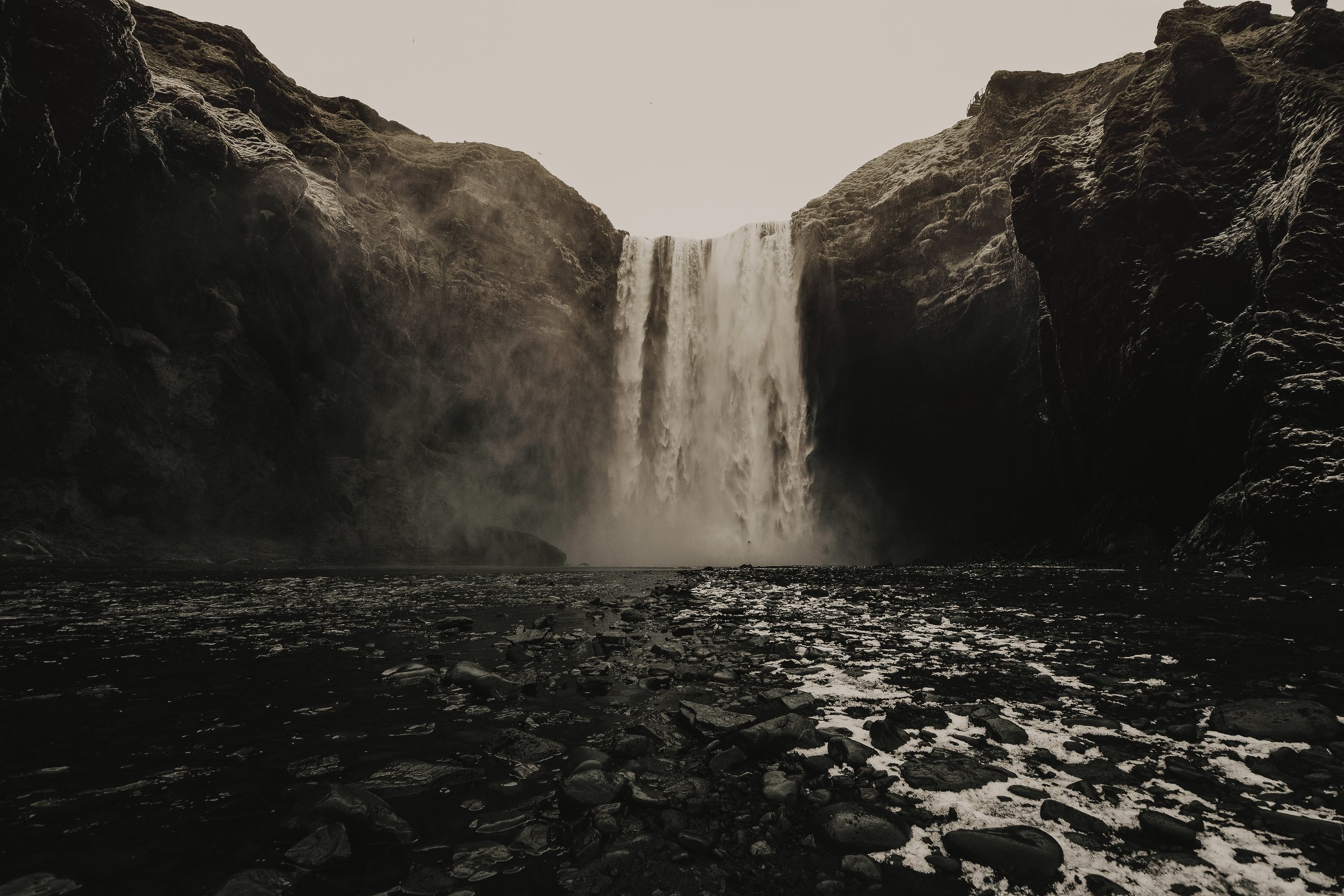 A waterfall in Iceland cascading down a rocky cliff into a river below, with dark rocks in the foreground and a pale sky above.