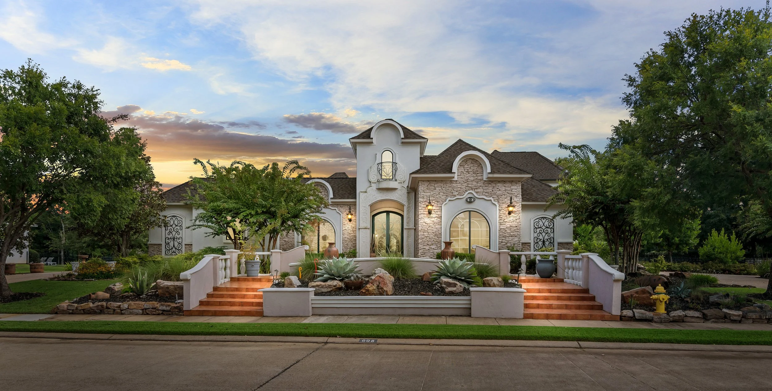 A house with a stone and stucco facade, arched windows, and a front porch with steps, surrounded by trees and landscaped yard at sunset.
