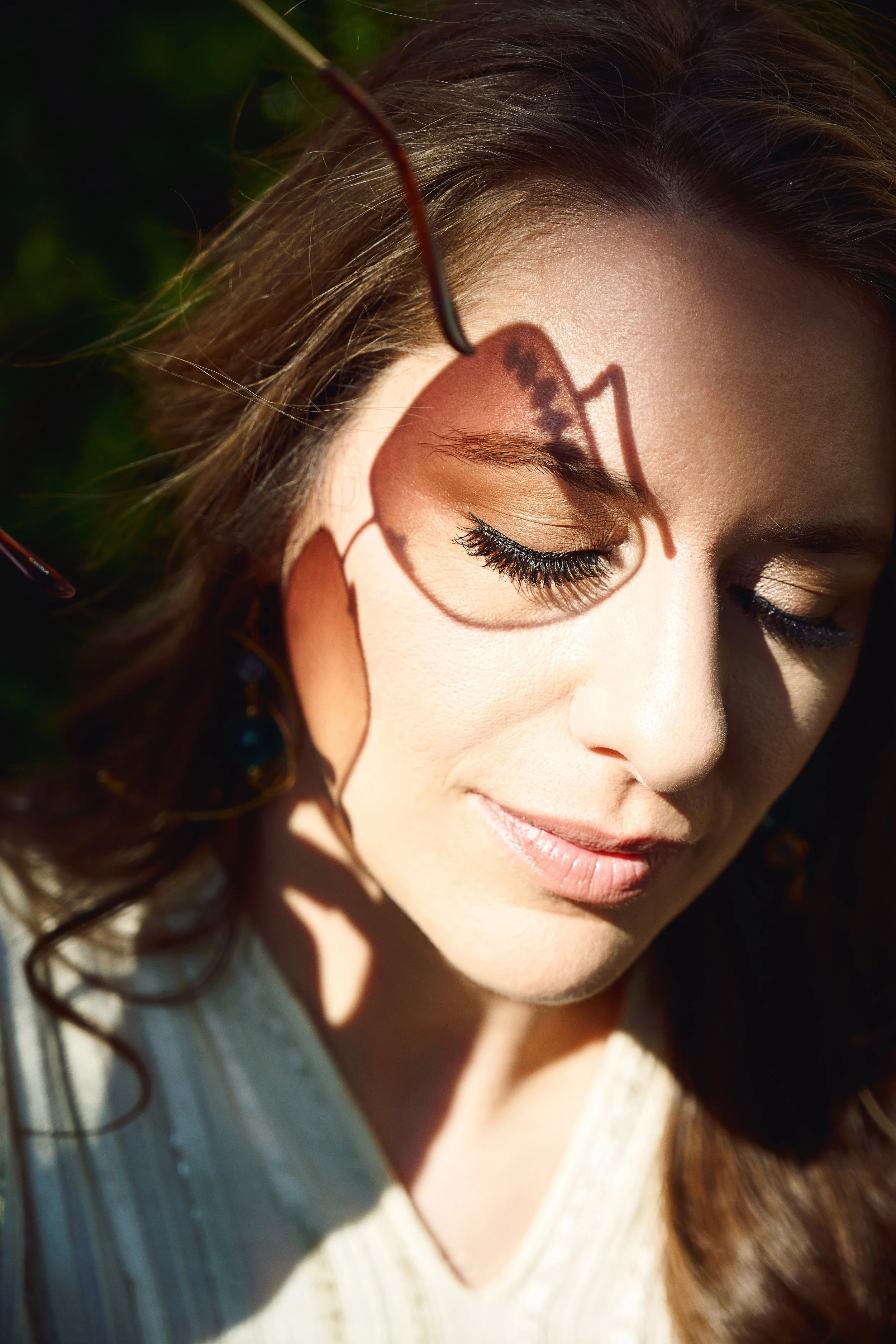 A woman with closed eyes, long eyelashes, and light makeup, sitting outdoors with sunlight casting a shadow of her glasses on her face.