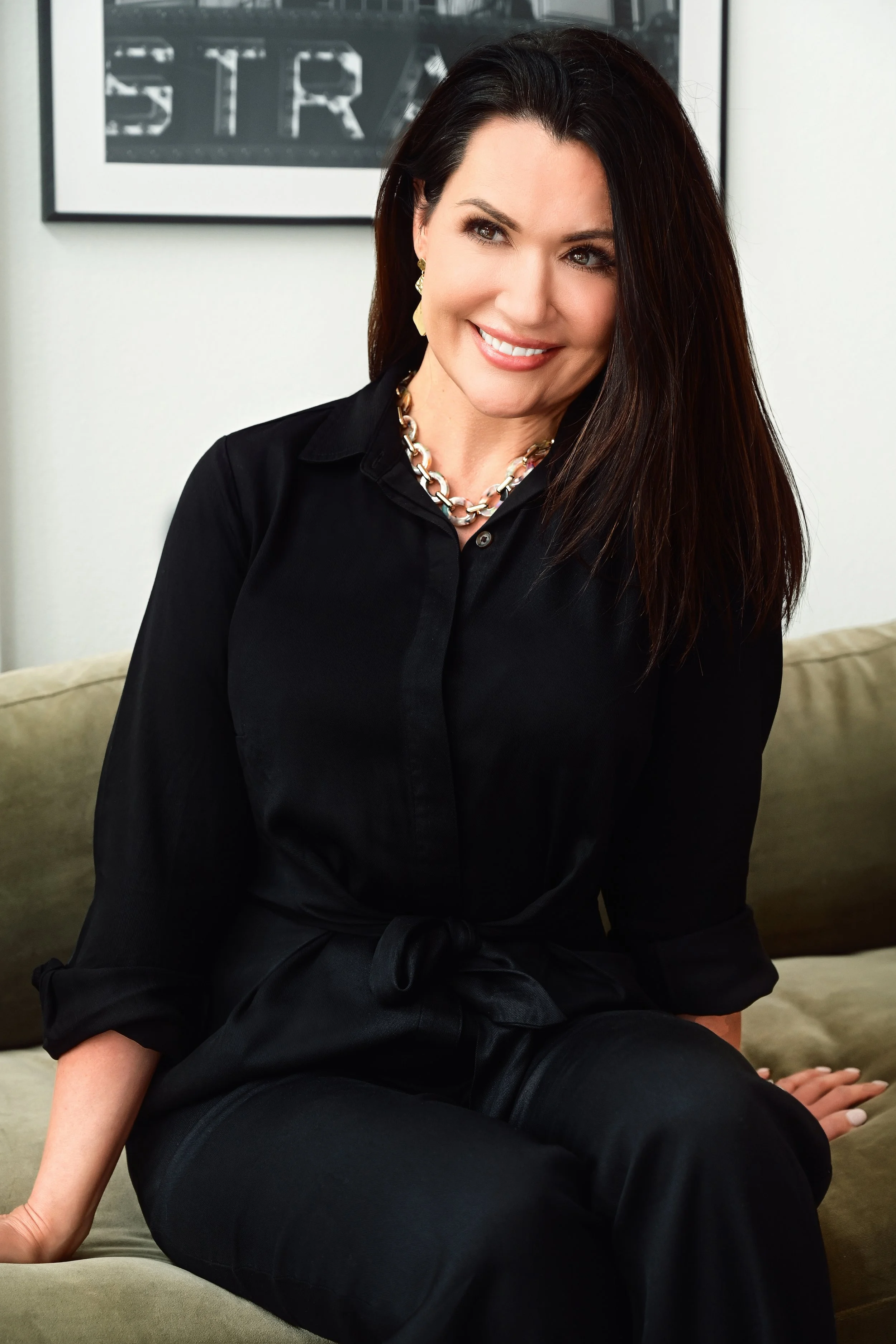 A woman with long dark hair, wearing a black shirt, gold earrings, and a chunky gold chain necklace, sitting on a beige couch, smiling.