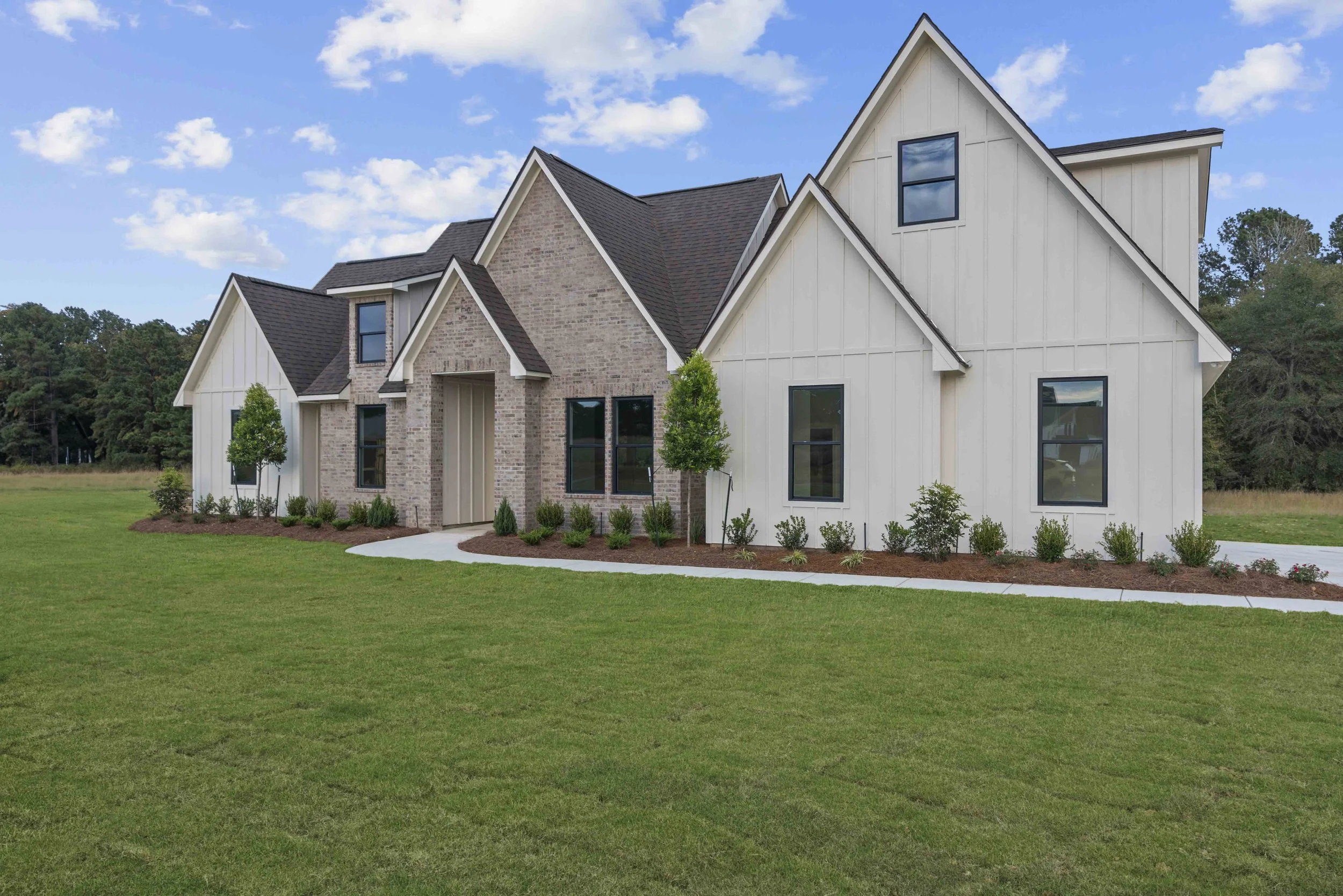 A modern house with a combination of brick and light-colored siding, multiple gabled roofs, and black-framed windows, surrounded by neatly landscaped lawn and small bushes under a partly cloudy sky.