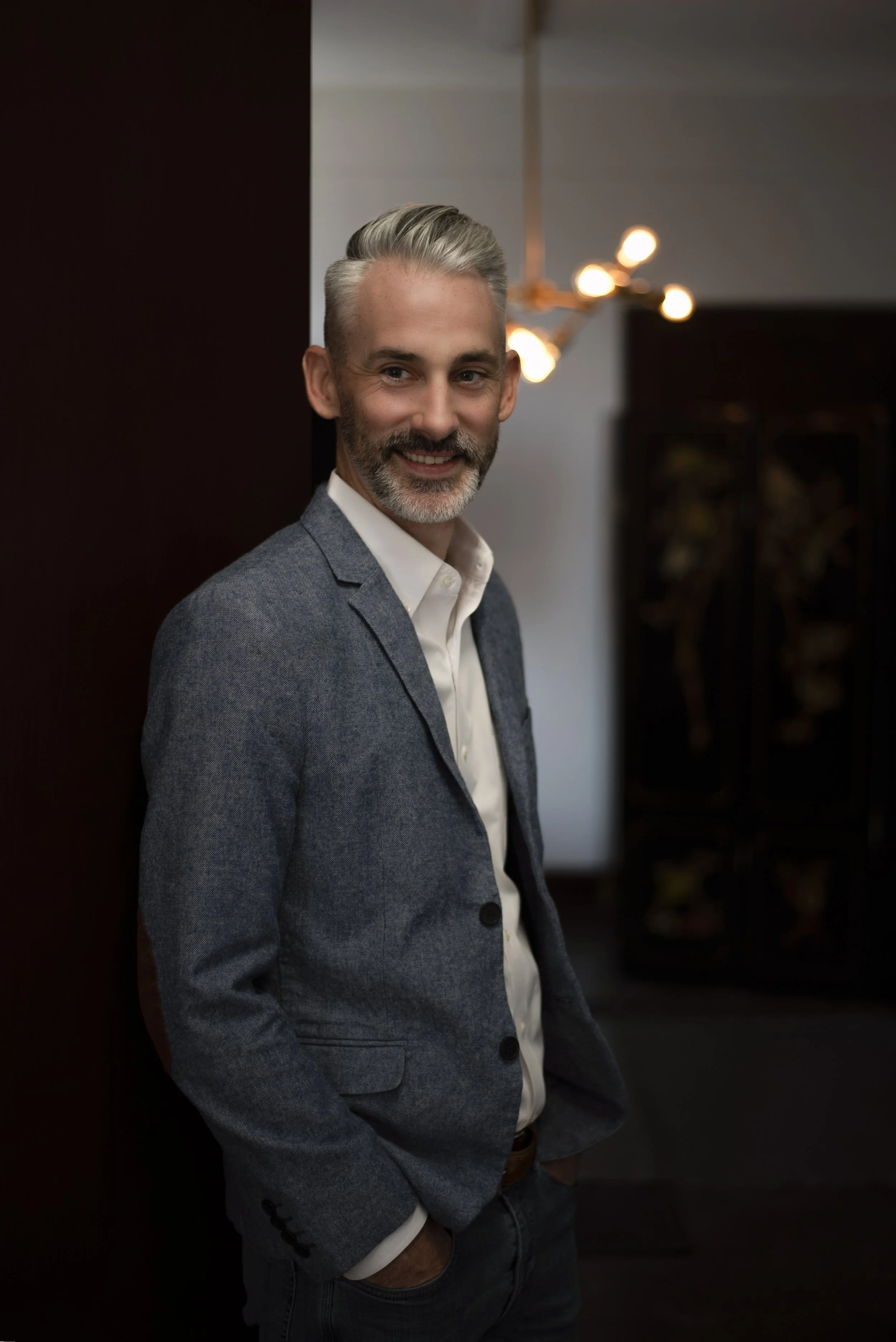 A smiling middle-aged man with gray hair and beard, dressed in a gray blazer, white shirt, and dark jeans, standing indoors near a dark wall with a modern chandelier in the background.