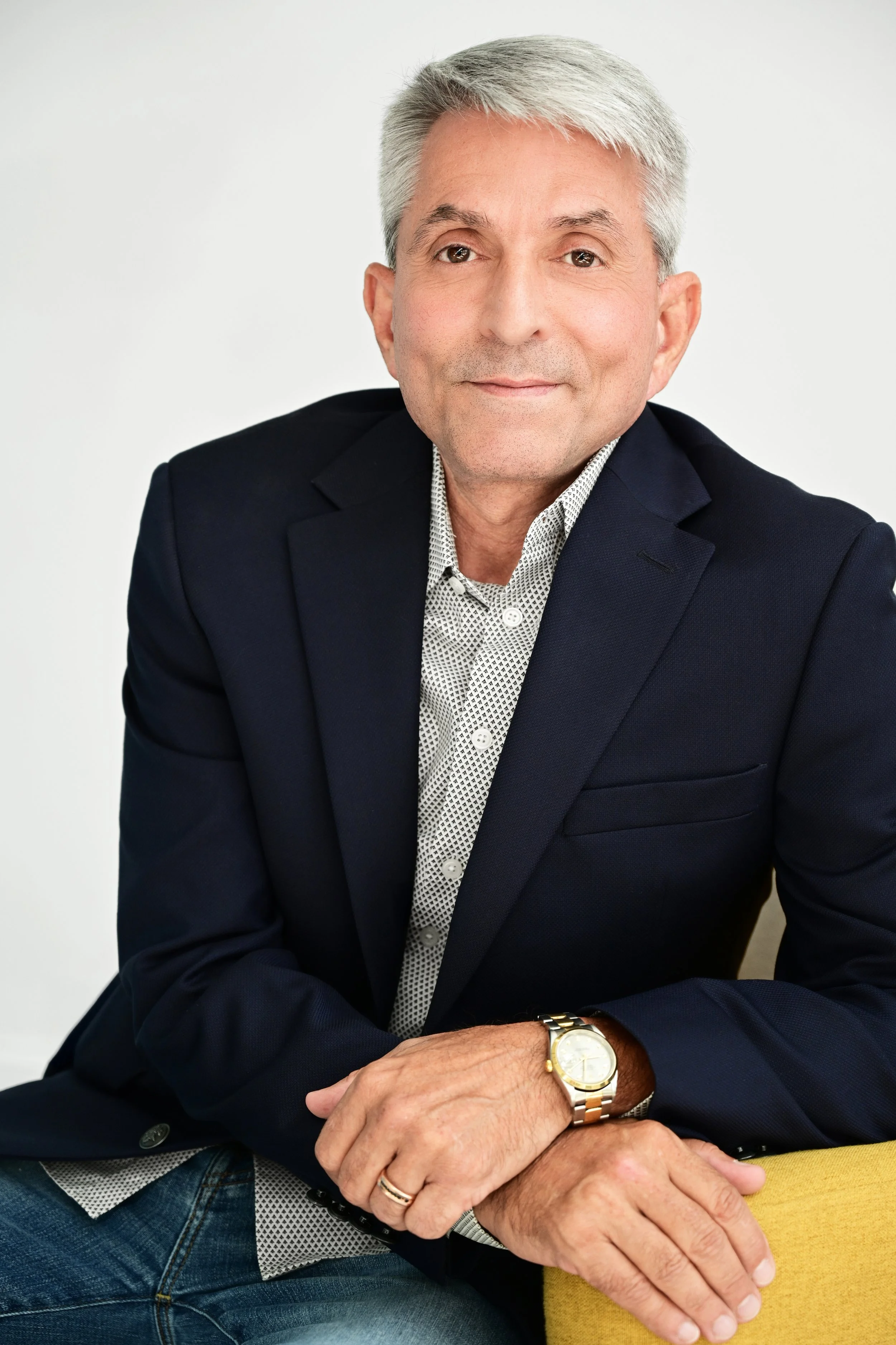 A middle-aged man with gray hair, dressed in a dark blazer and patterned shirt, is sitting with crossed arms, wearing a gold watch, and smiling slightly at the camera. The background is plain white.