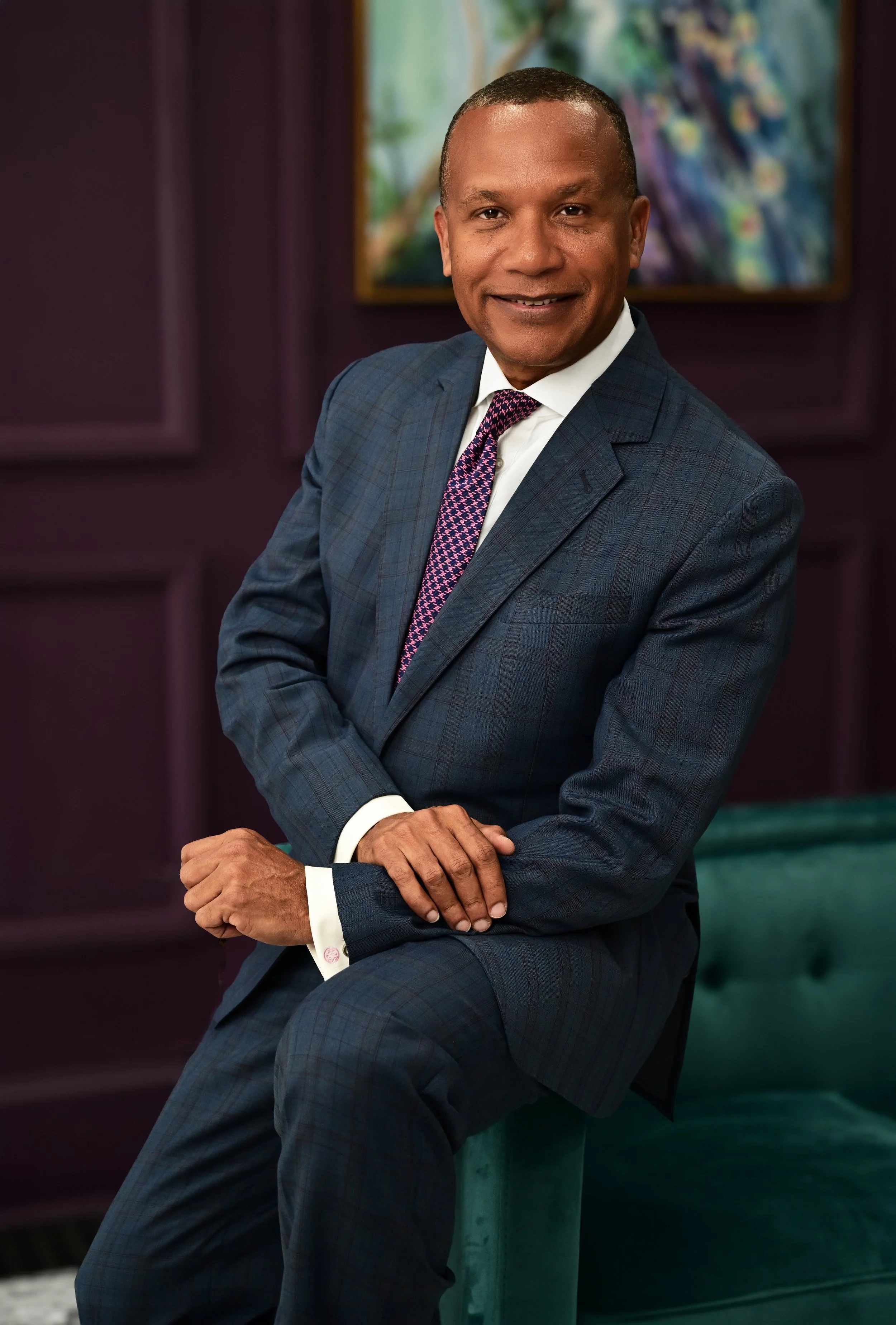 A man in a blue checkered suit with a pink patterned tie, sitting on a ledge with his right arm resting on his knee, smiling at the camera, with a framed painting of a nature scene in the background.