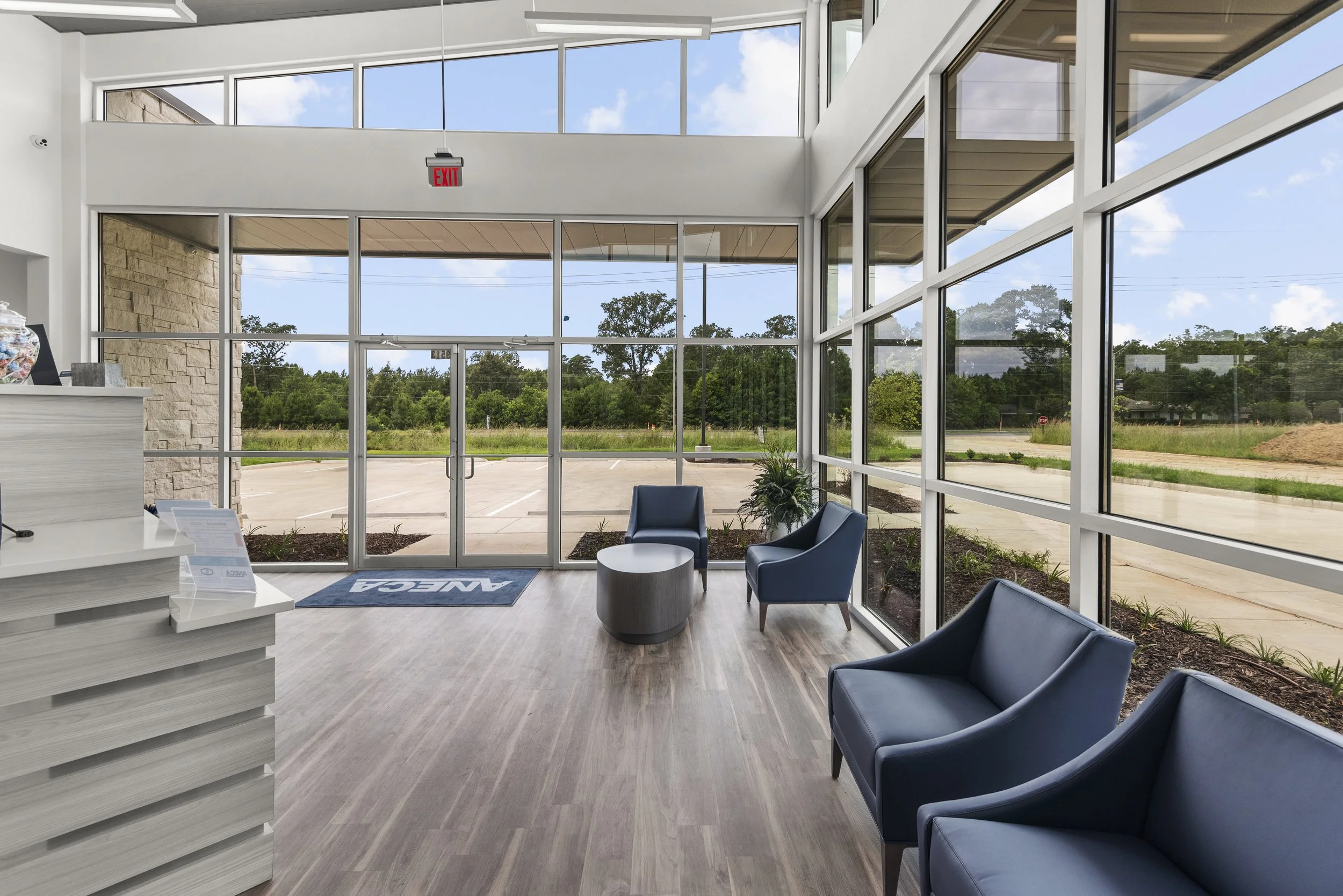 Modern lobby with large glass windows, blue chairs, and potted plants, overlooking a parking lot and trees outside.