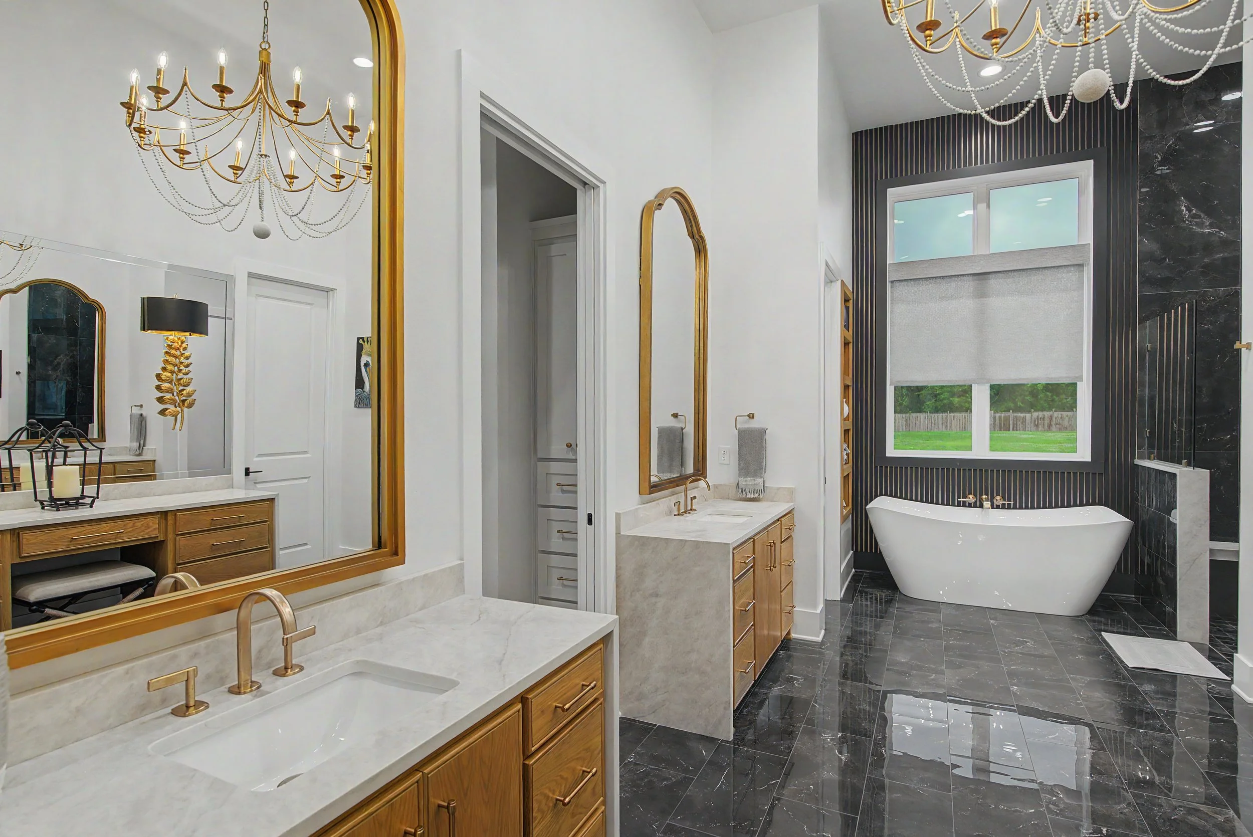 A modern bathroom with a large bathtub, black and white marble flooring, a black marble wall, gold fixtures, a vanity with a white marble countertop, a gold-framed mirror, a black and gold lampshade, decorative chandeliers, and a window showing green