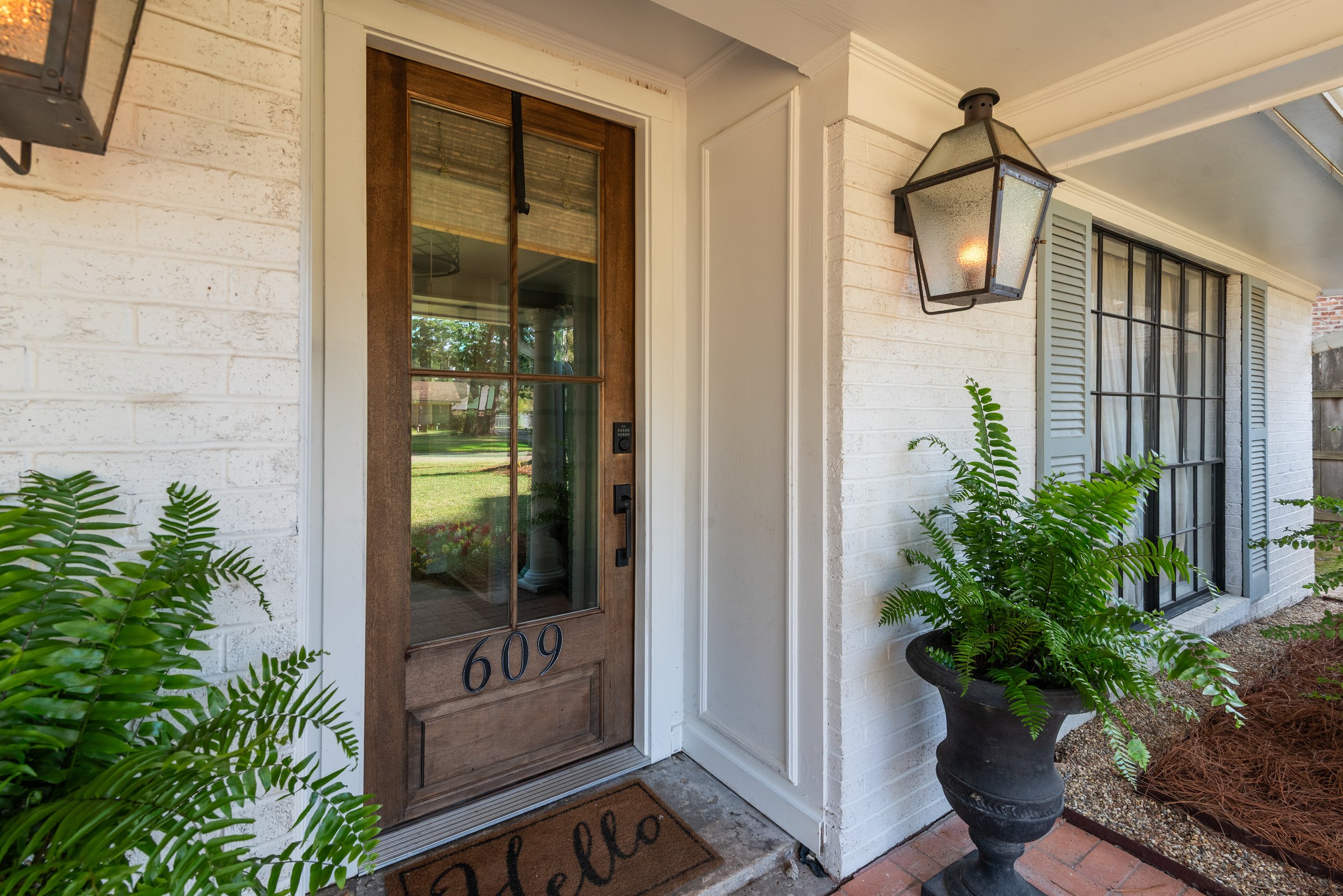 Front door of a house with a wooden frame, glass panels, and the number 609. Potted fern plant in a large black pot is on the right side. Light fixture above the door and a welcome mat in front.