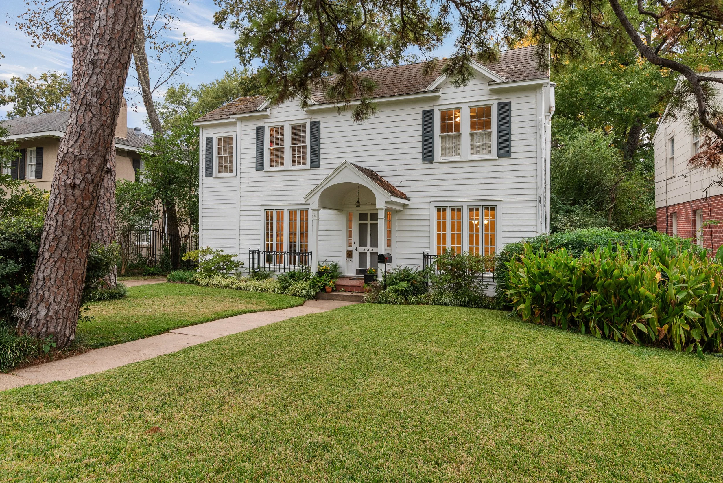 White two-story house with black shutters, front porch, and green lawn, surrounded by trees and shrubs.