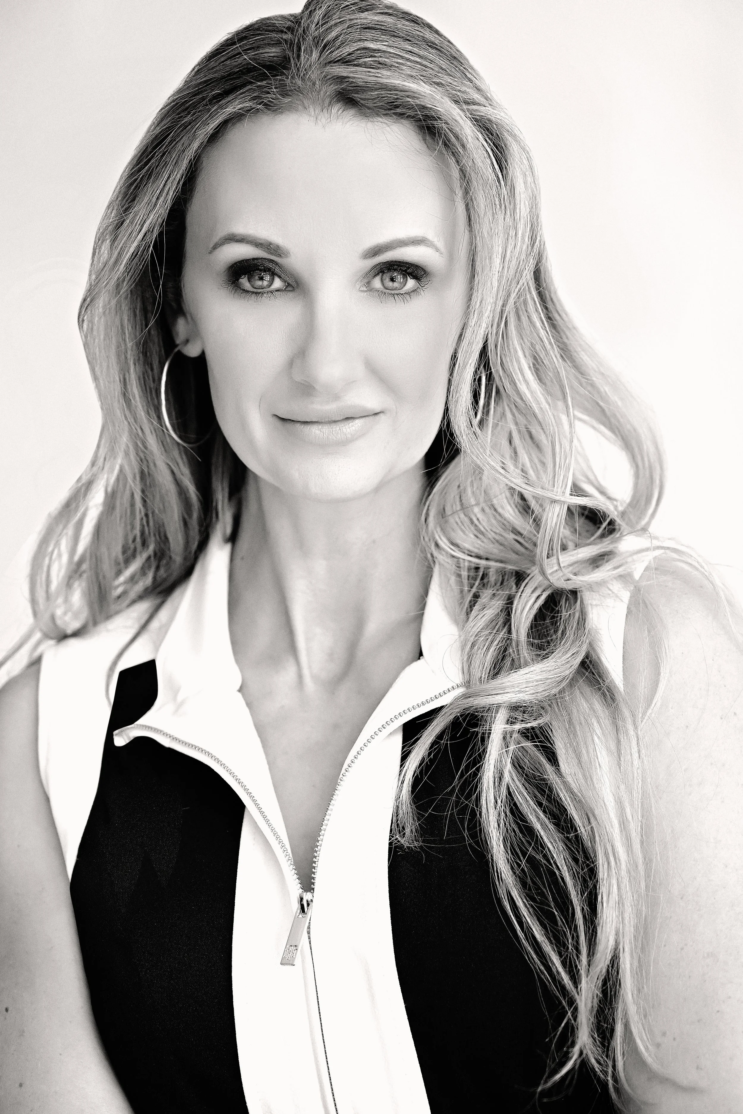 Black and white portrait of a woman with long wavy hair, wearing a sleeveless zip-up athletic top with a black and white pattern, and large hoop earrings.